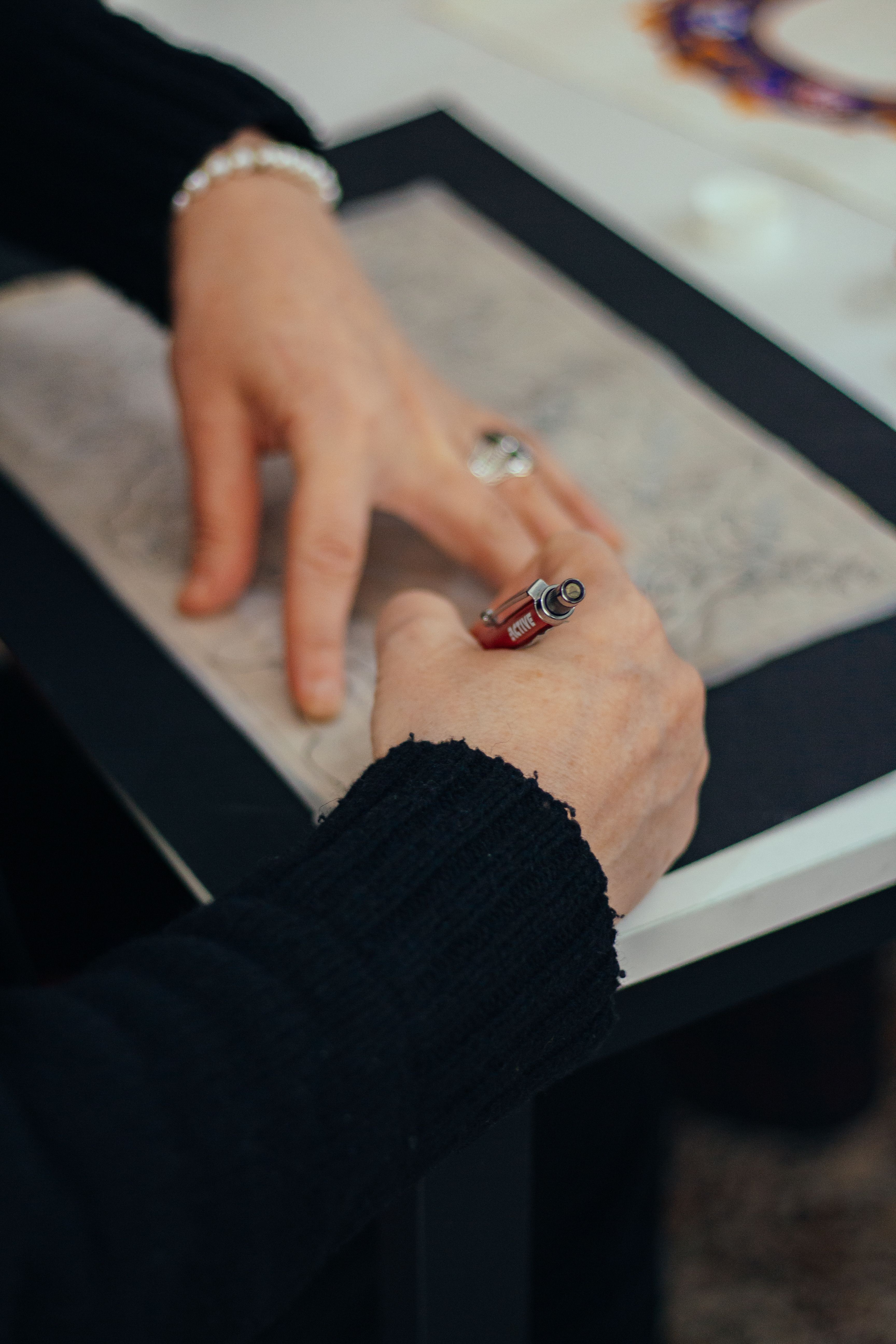 Close-up of hands sketching on paper with a red mechanical pencil, surrounded by a dark drawing board and blurred supplies.