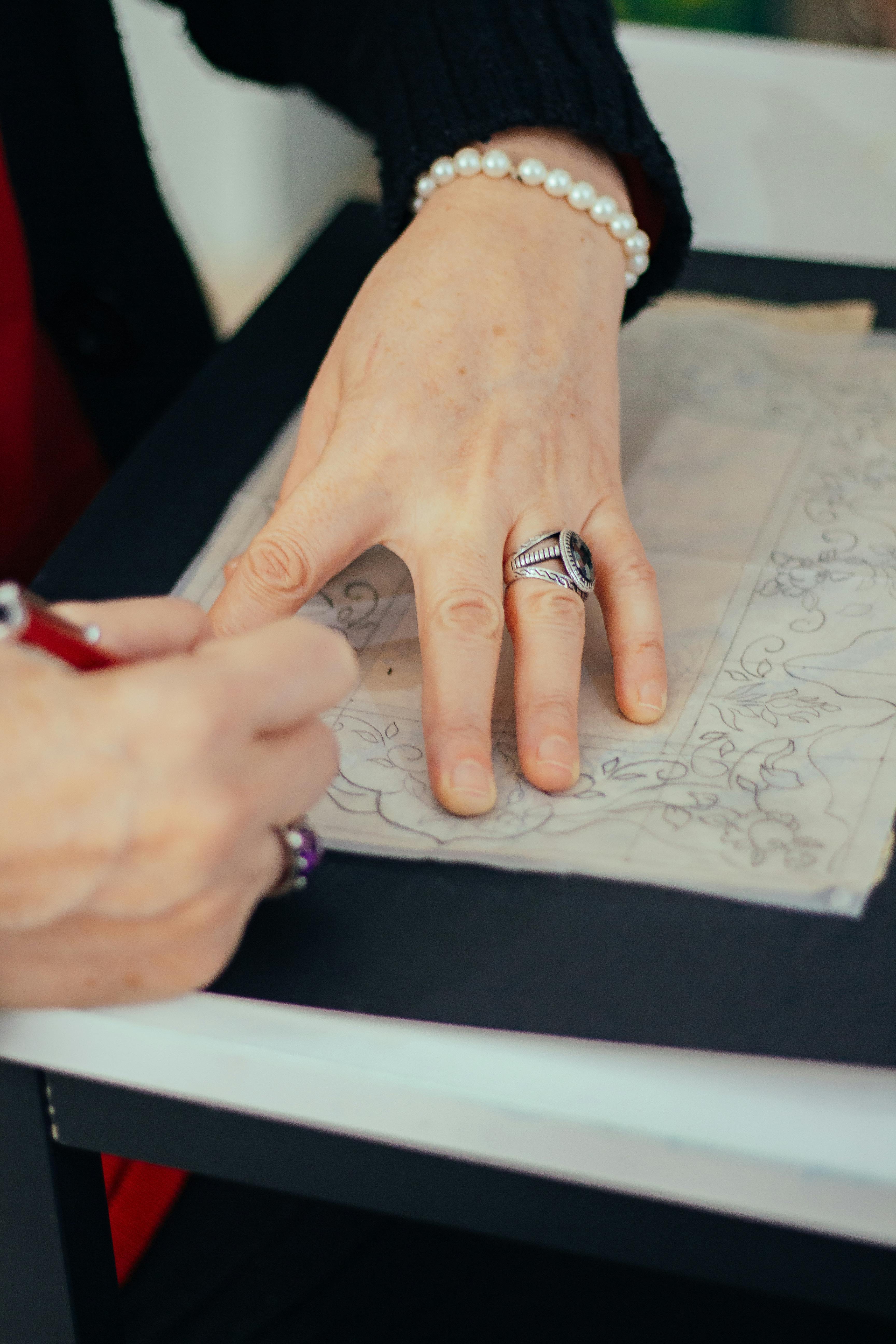 Close-up of a person sketching an intricate design on paper, wearing pearl bracelet and rings.