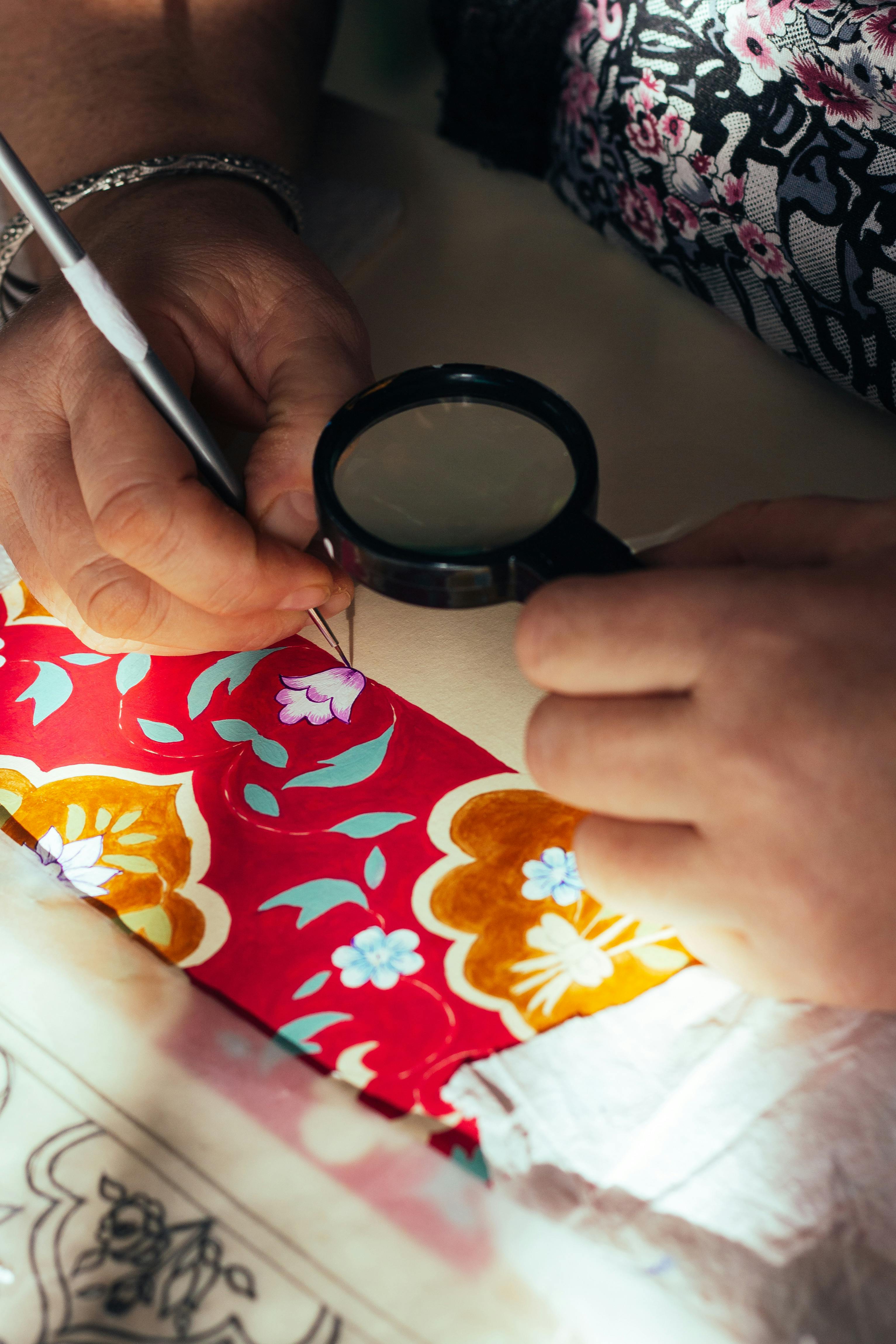 Detailed view of hands examining vibrant patterned cloth with magnifying glass.