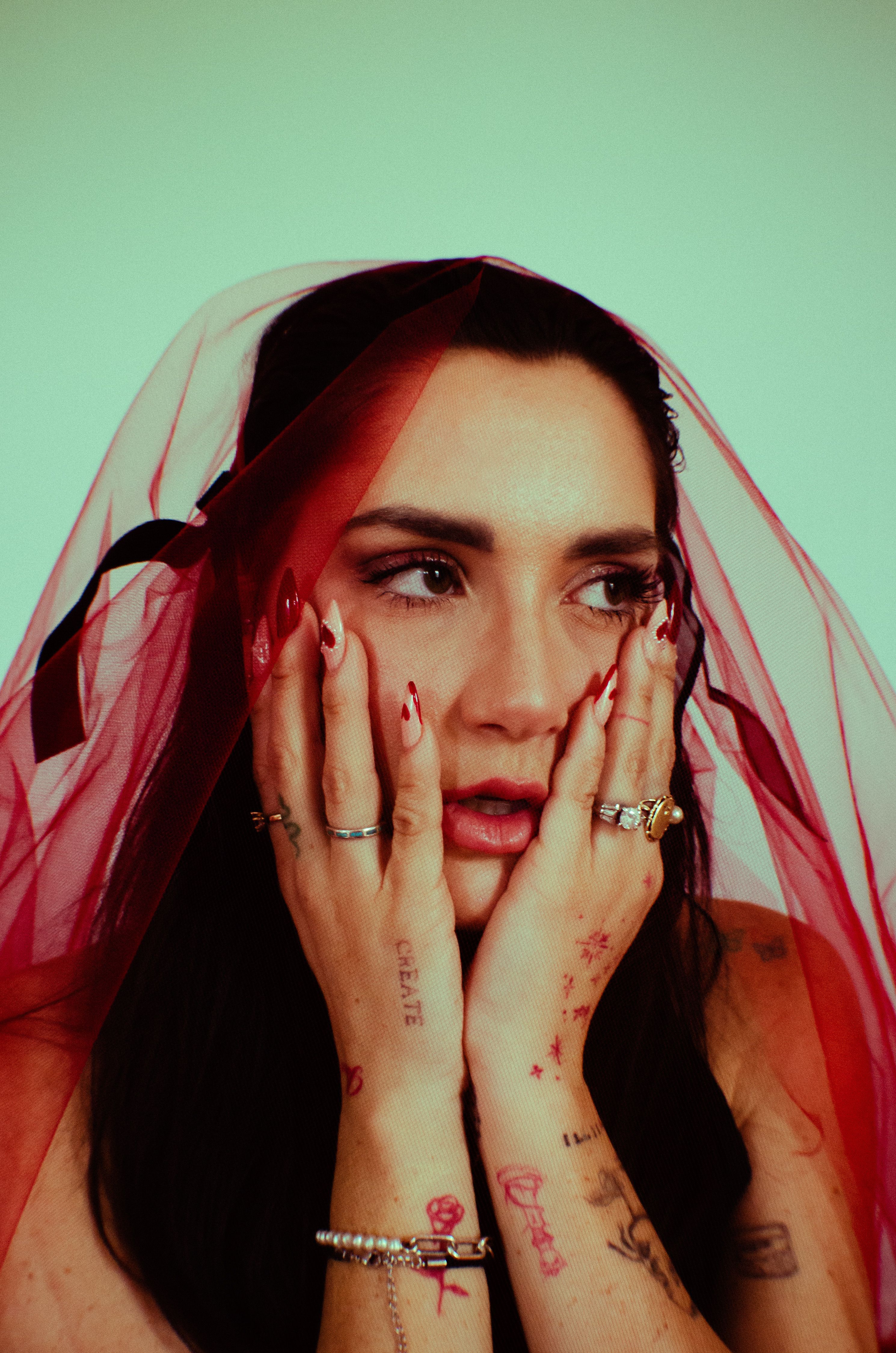 Portrait of a thoughtful woman wearing a red veil, showcasing tattoos and rings.