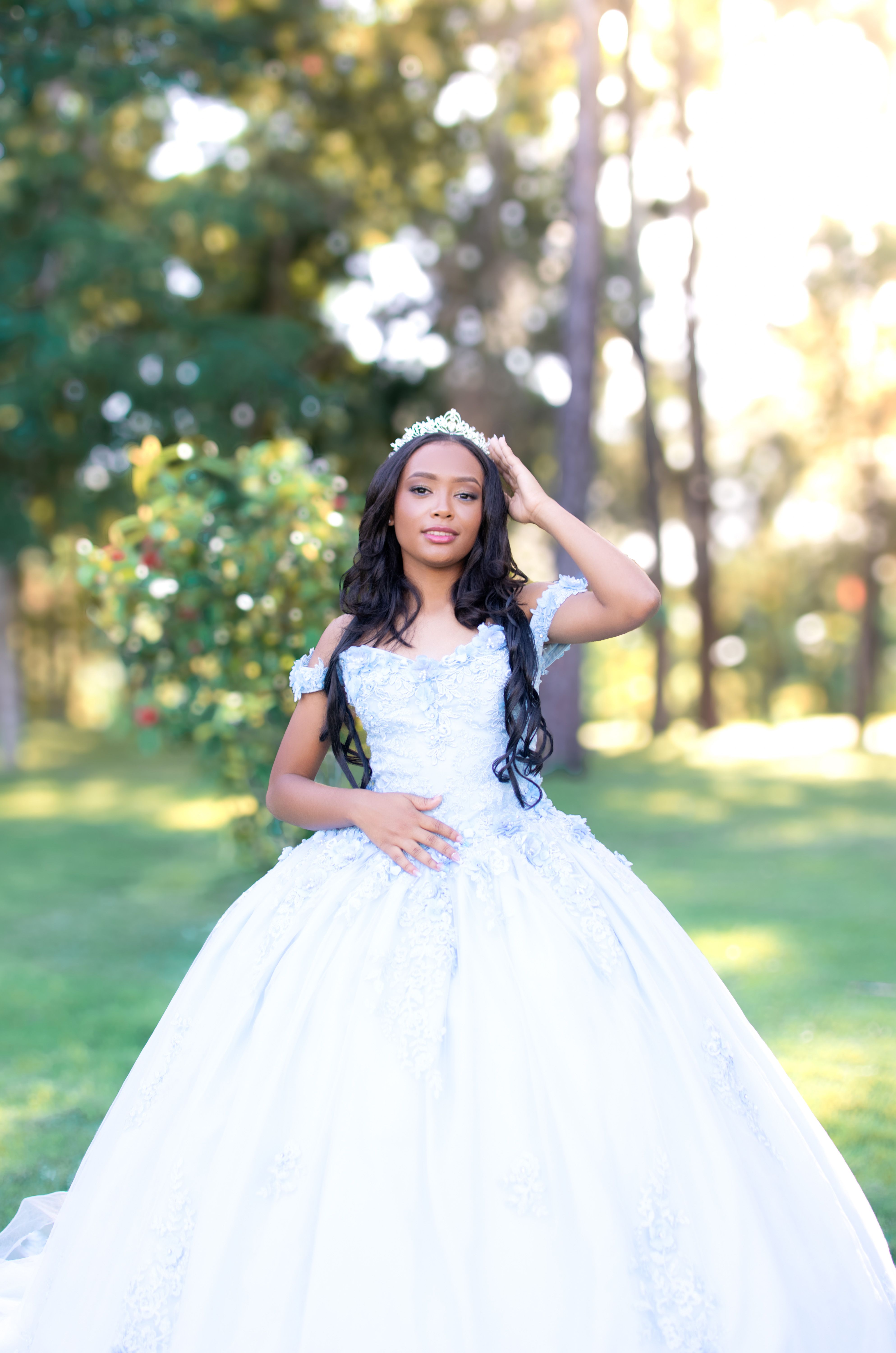 Young woman in a beautiful white quinceañera gown posing outdoors in a sunlit garden.