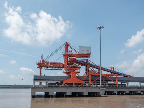 A large orange industrial crane situated at a waterside port under a blue sky.