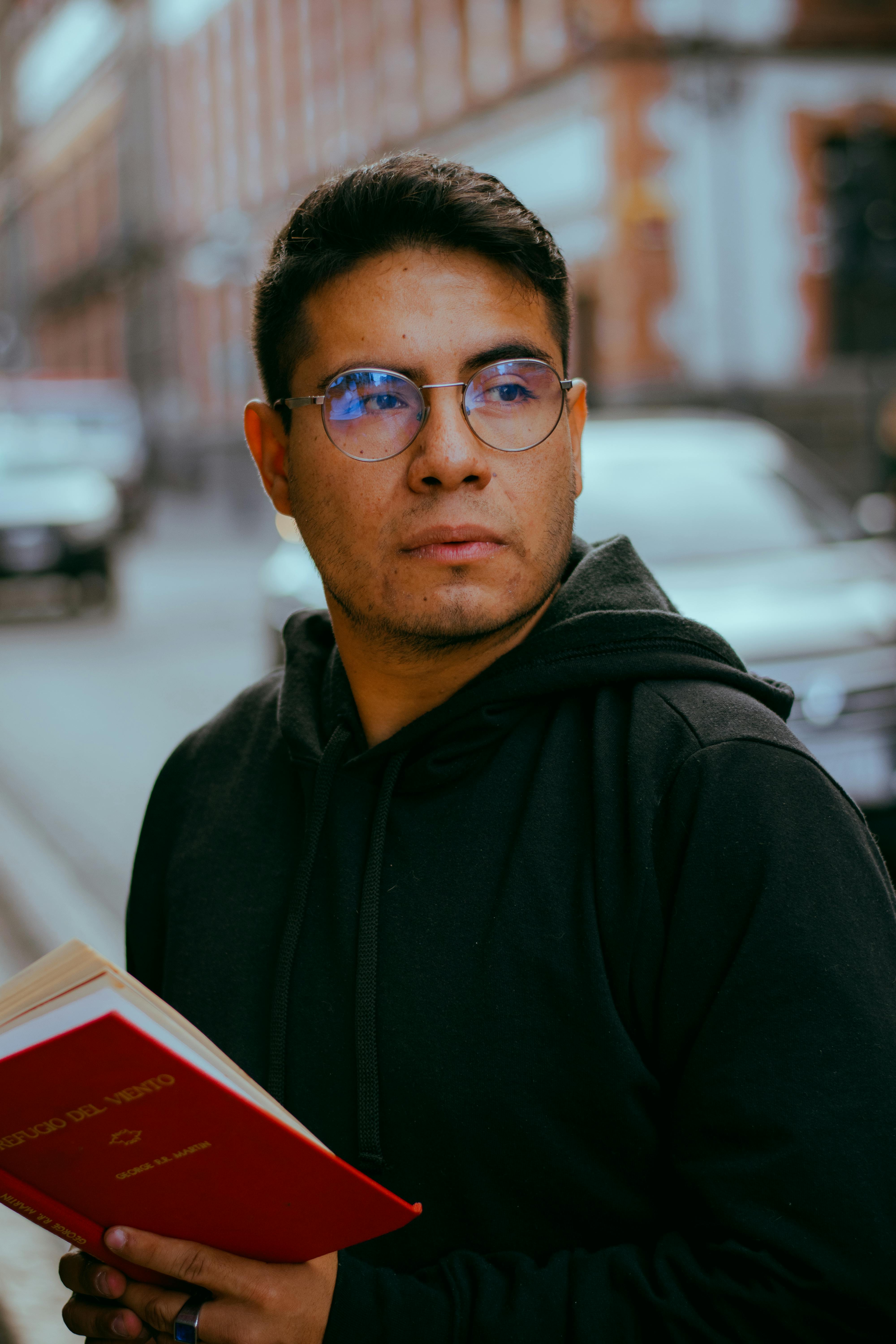 Young man with glasses reading a book outdoors on an urban street during the day.