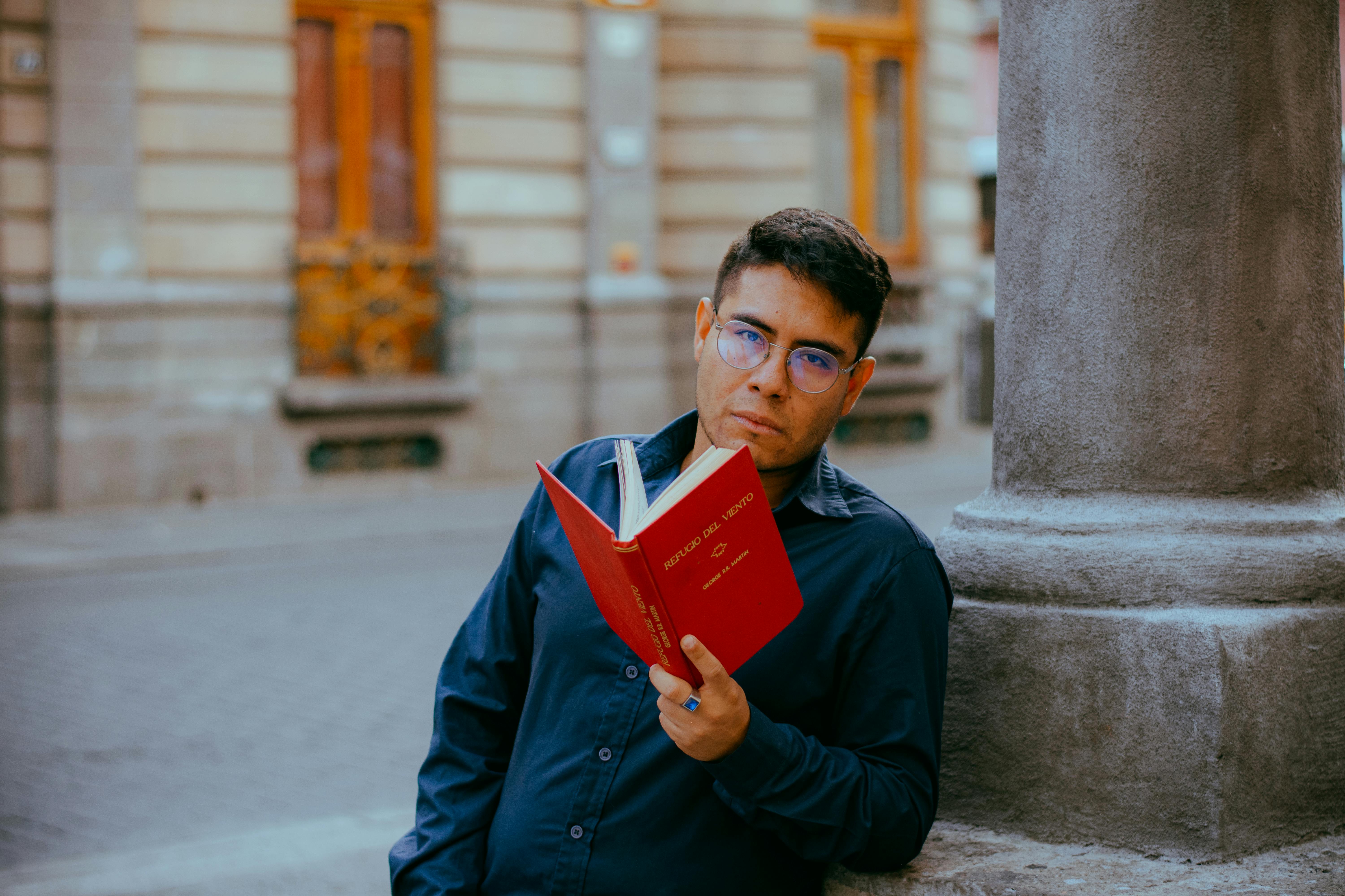 A man in glasses reading a red book against a stone column in a city.
