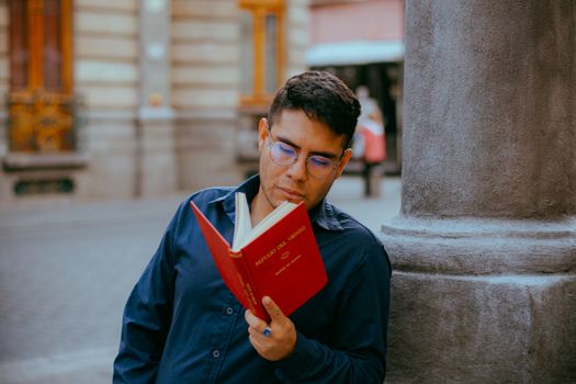 A man in glasses reading a red book while leaning against a column in a city street.