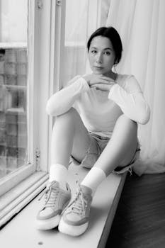 Thoughtful young woman sitting by a window in casual attire, in black and white.