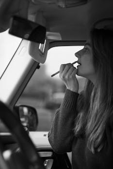 Black and white image of a woman putting on lipstick in a car interior.