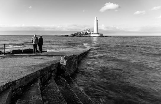 Black and white seascape with lighthouse and people on pier.