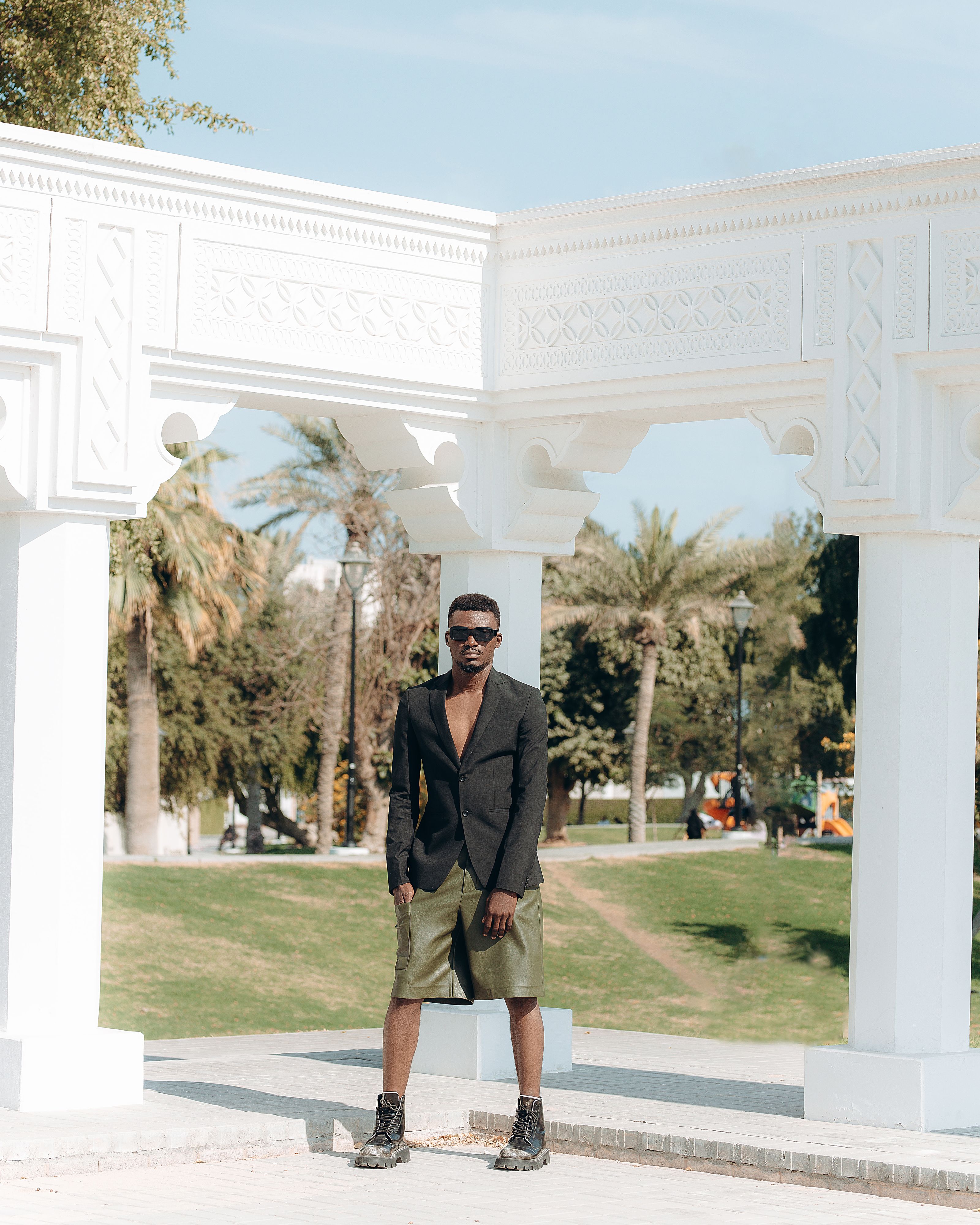 Homem elegante de óculos escuros posando sob um arco branco em um parque.