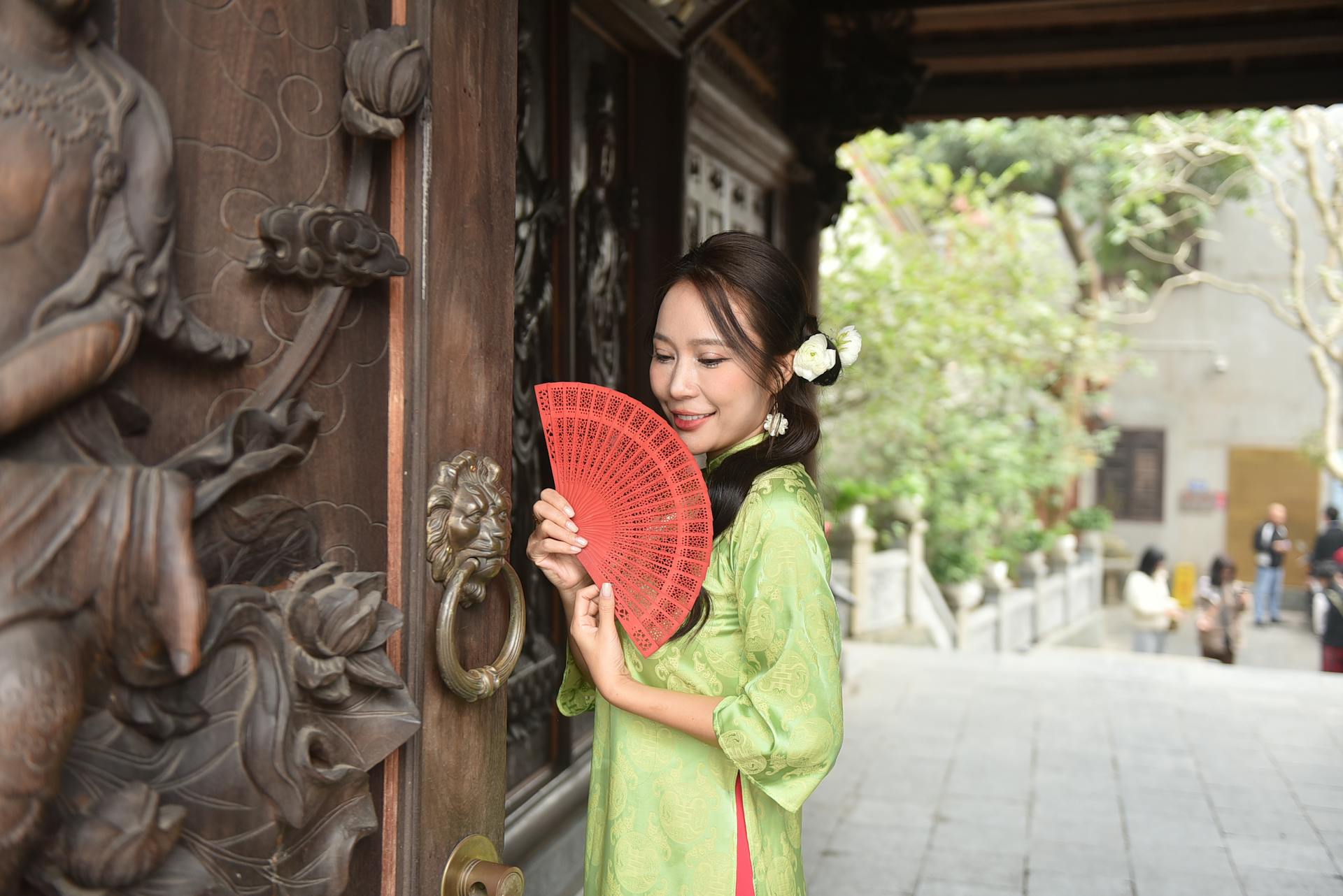 Asian woman in traditional dress holding a red fan outside a temple with intricate wood carvings.