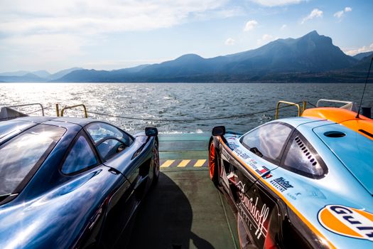 Supercars parked by Lake Garda with stunning mountains in the background, under a sunny sky.