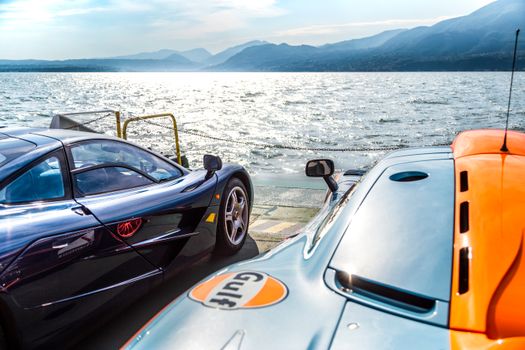 Two stunning supercars parked by Lake Garda with a breathtaking mountainous backdrop.