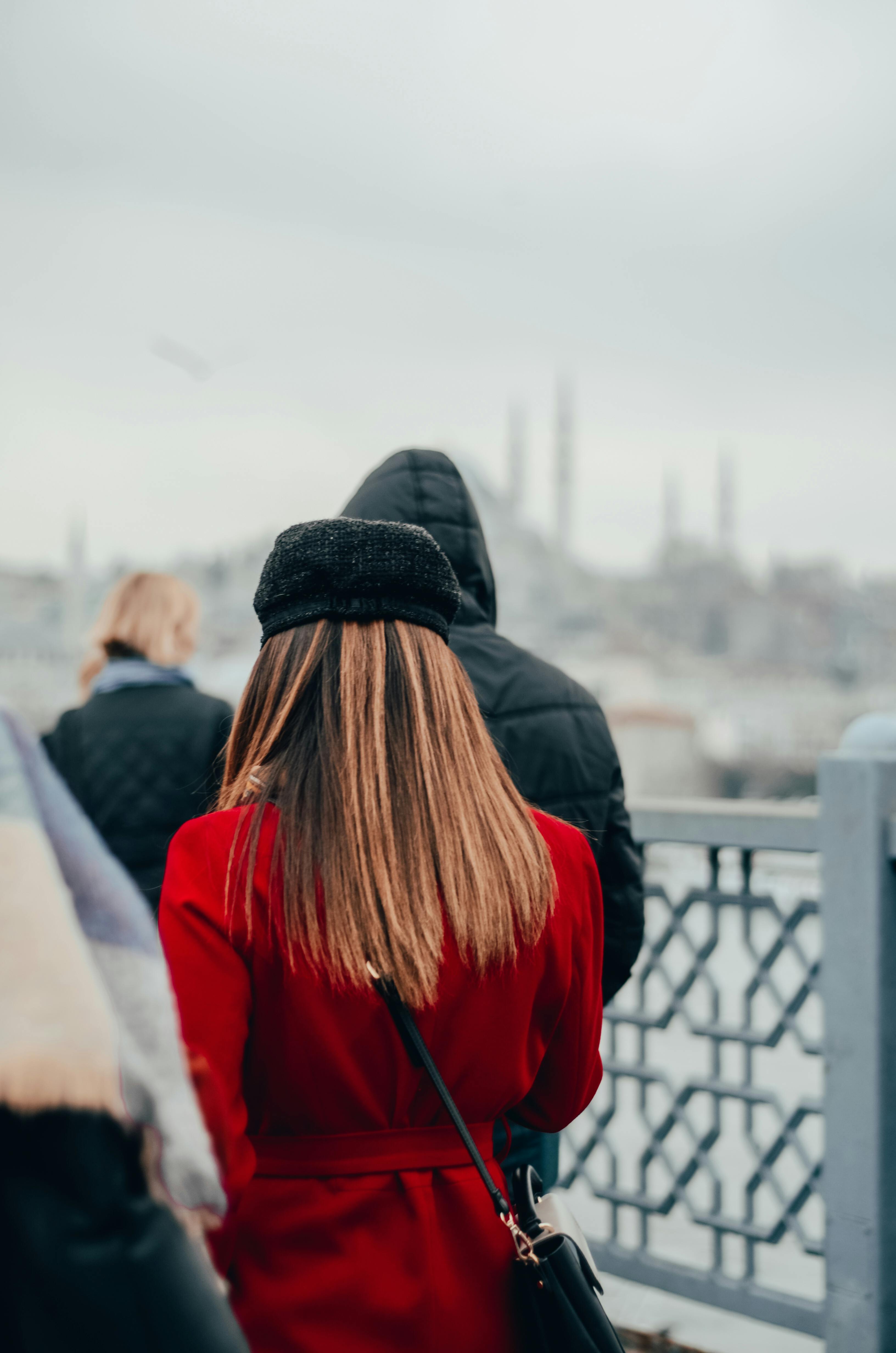一位穿着红色外套、戴着帽子的时尚女性，在冬日里走过城市天桥，背景是模糊的城市景观