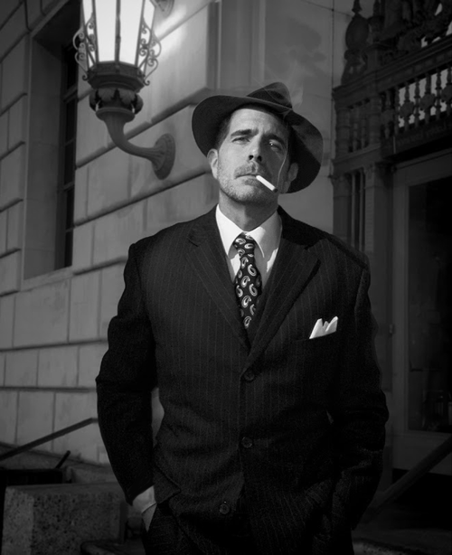 Free A man in a vintage suit and fedora exudes film noir vibes, smoking outdoors. Stock Photo