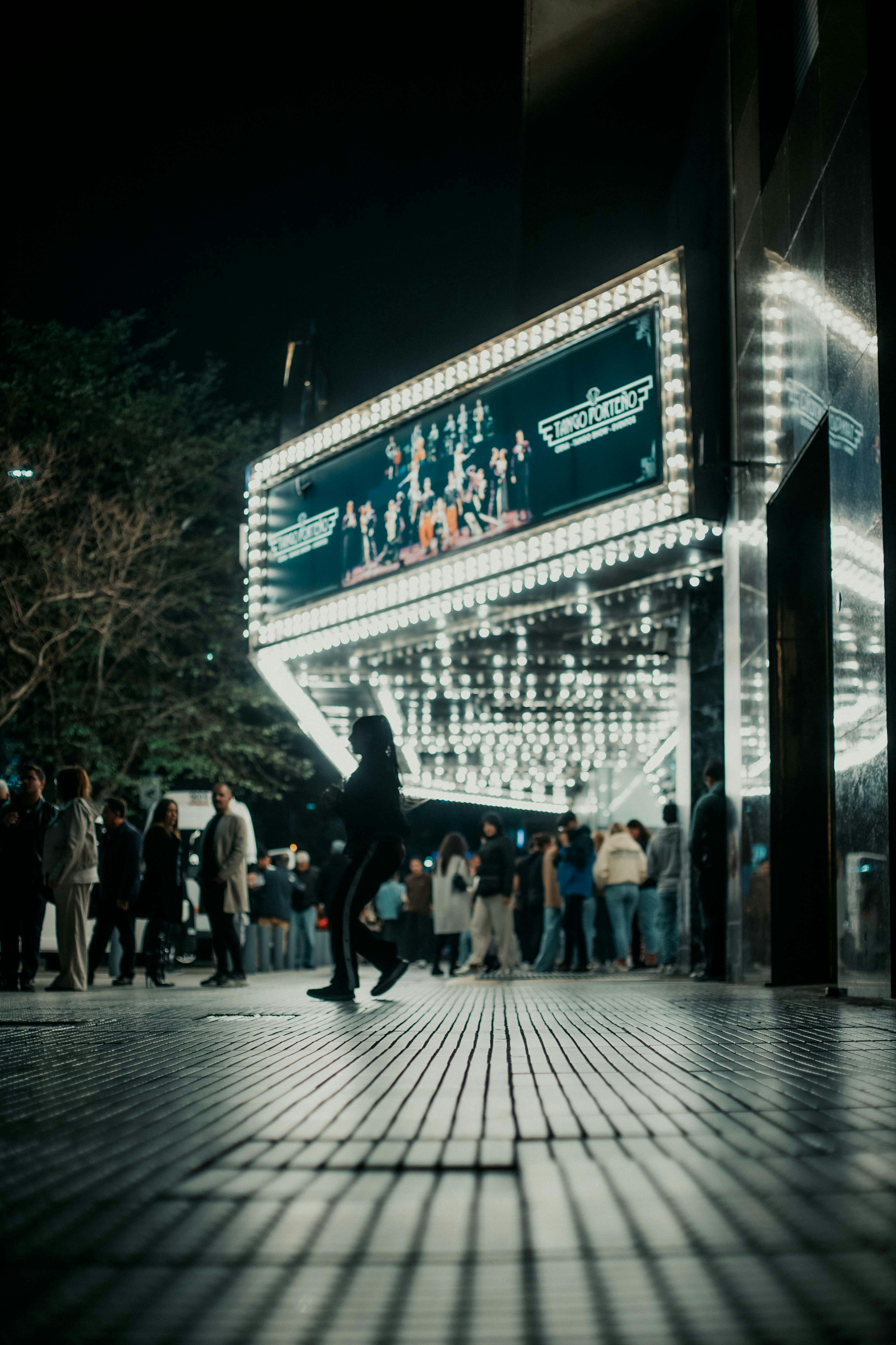 grátis Pessoas em silhueta caminham por um teatro bem iluminado com uma marquise à noite. Foto profissional