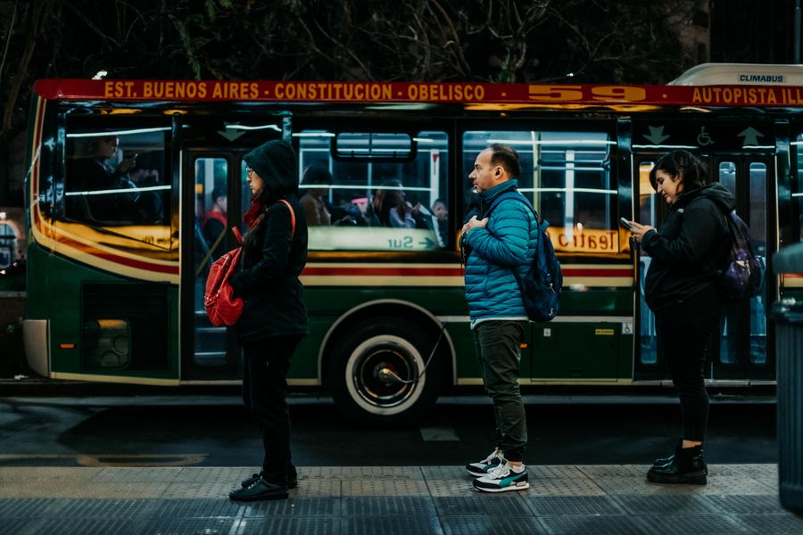 Commuters line up at night in Buenos Aires, waiting beside a colorful city bus.