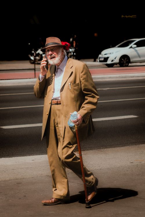 Free Senior man in a brown suit walking on a city street, using a cane and talking on a phone. Stock Photo