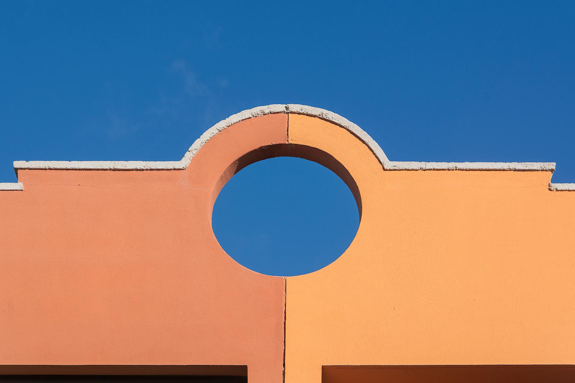 Bright facade with circular cutout contrasting a clear blue sky, showcasing architectural design.