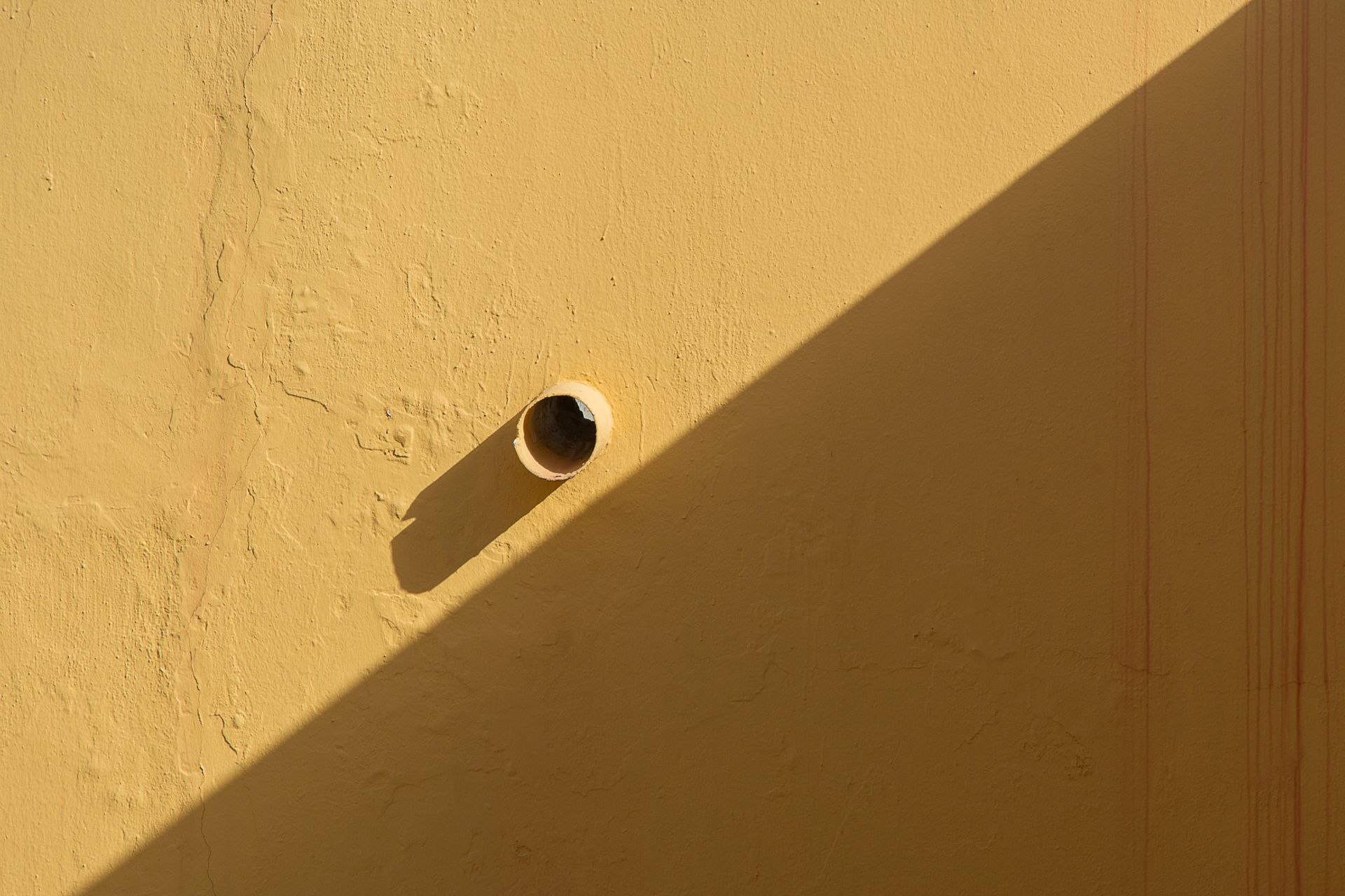 A yellow wall featuring a protruding pipe and strong geometric shadow cast in natural light.