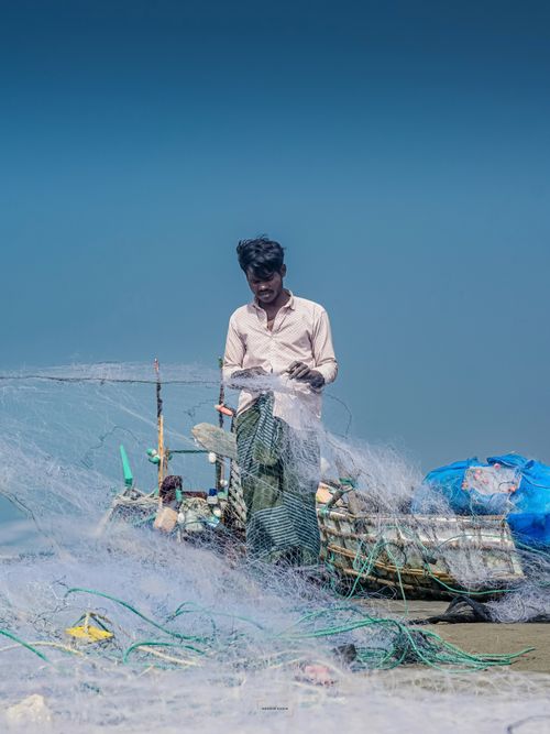 Free A fisherman repairs his net aboard a wooden boat, under clear blue skies, showcasing rural life. Stock Photo