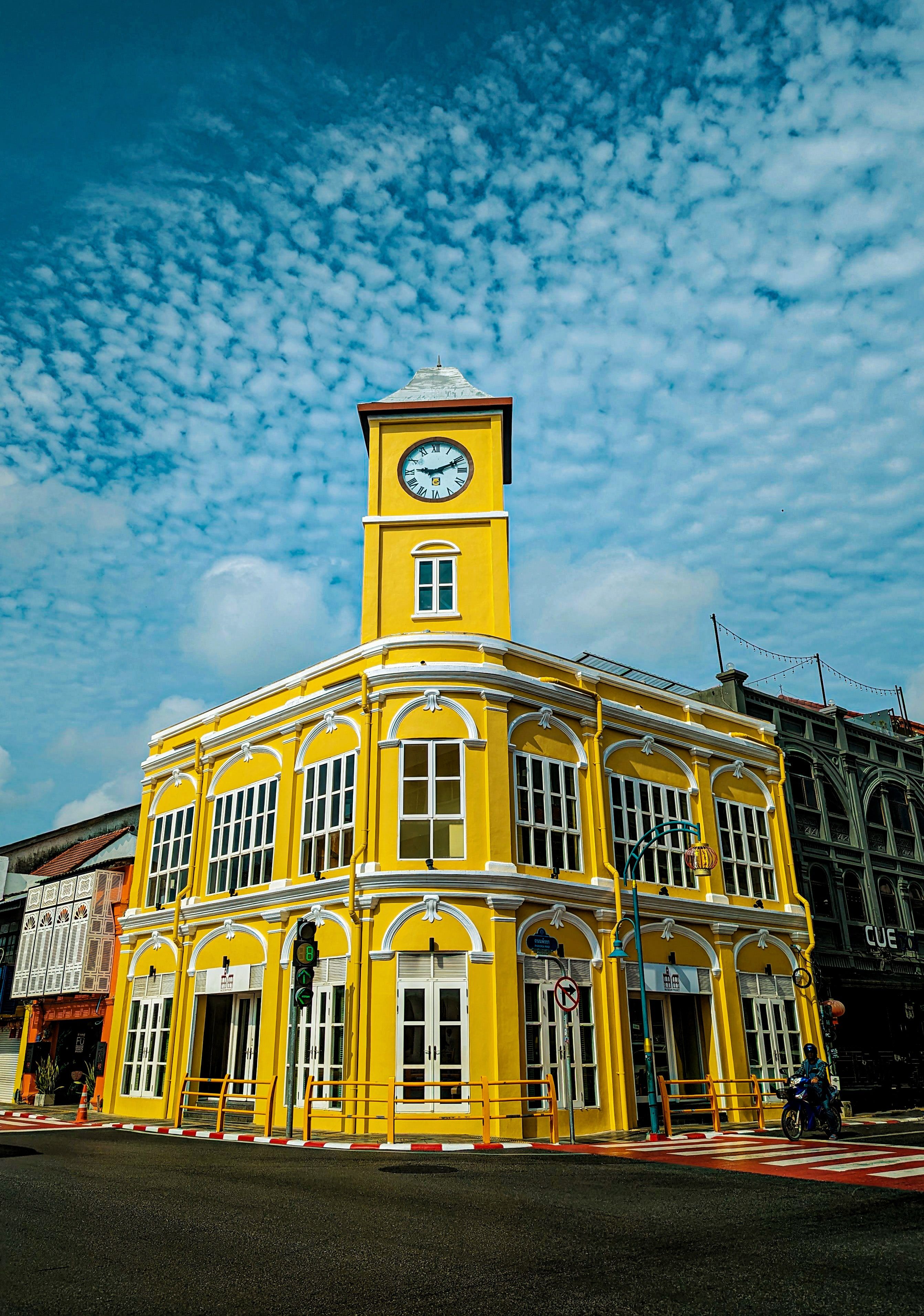 Ancient buildings in Sino-European style in Phuket, Thailand, captured as a bright yellow colonial corner building with clock tower under a textured blue sky.