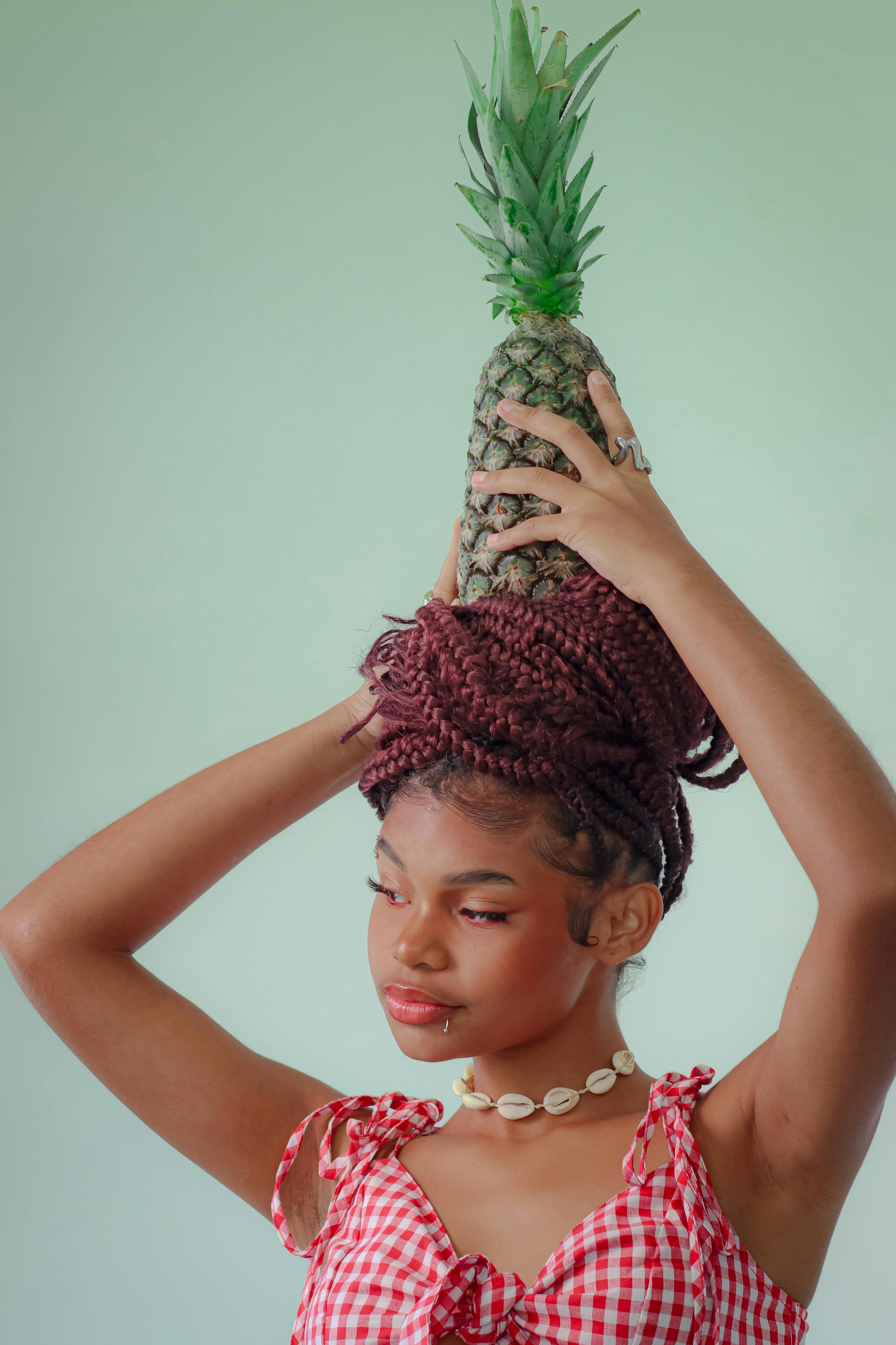 Stylish woman with braided hair holds a pineapple on her head, exuding a playful, quirky vibe.