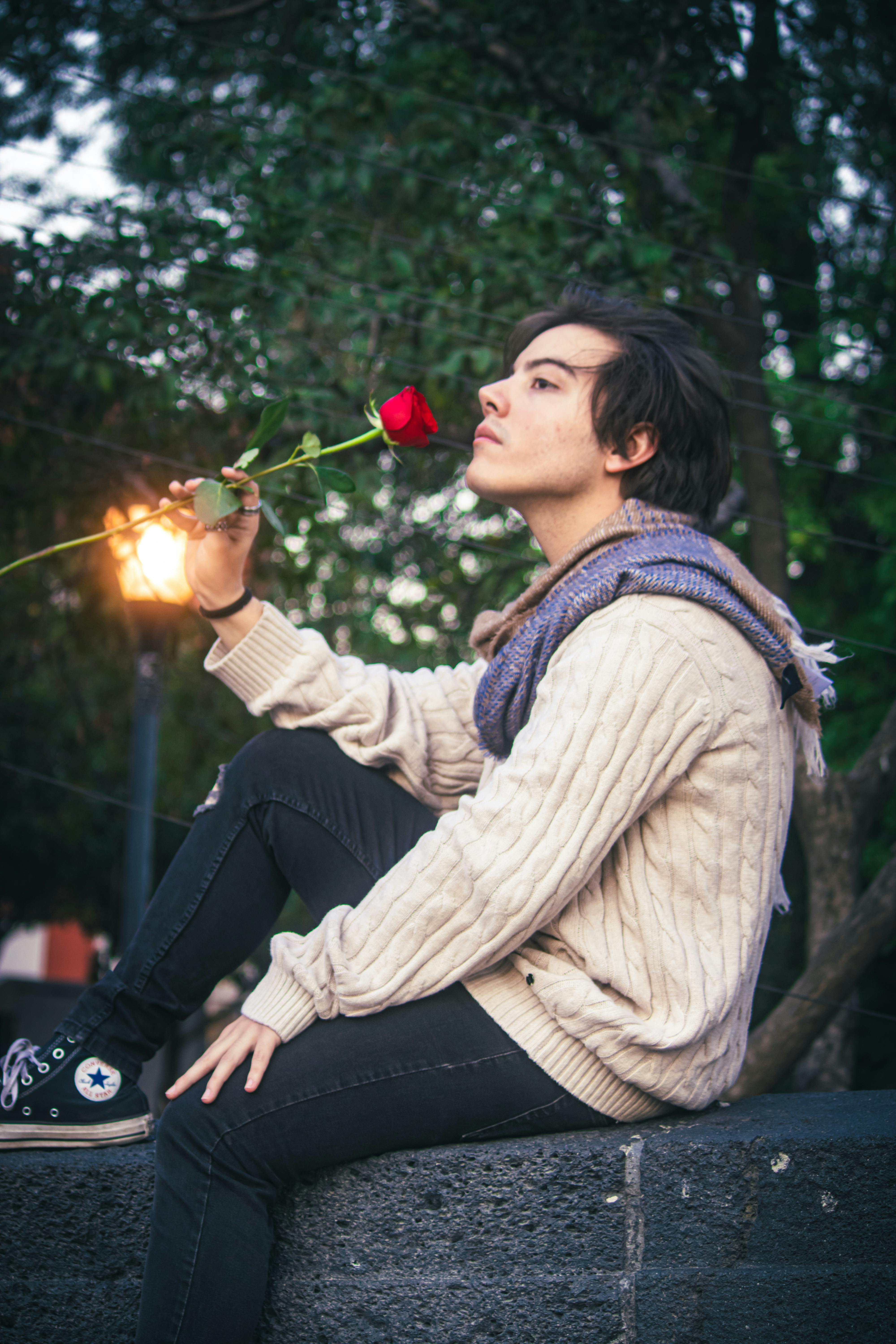 A young man holding a red rose, sitting outdoors in a park with a romantic vibe in the evening light.