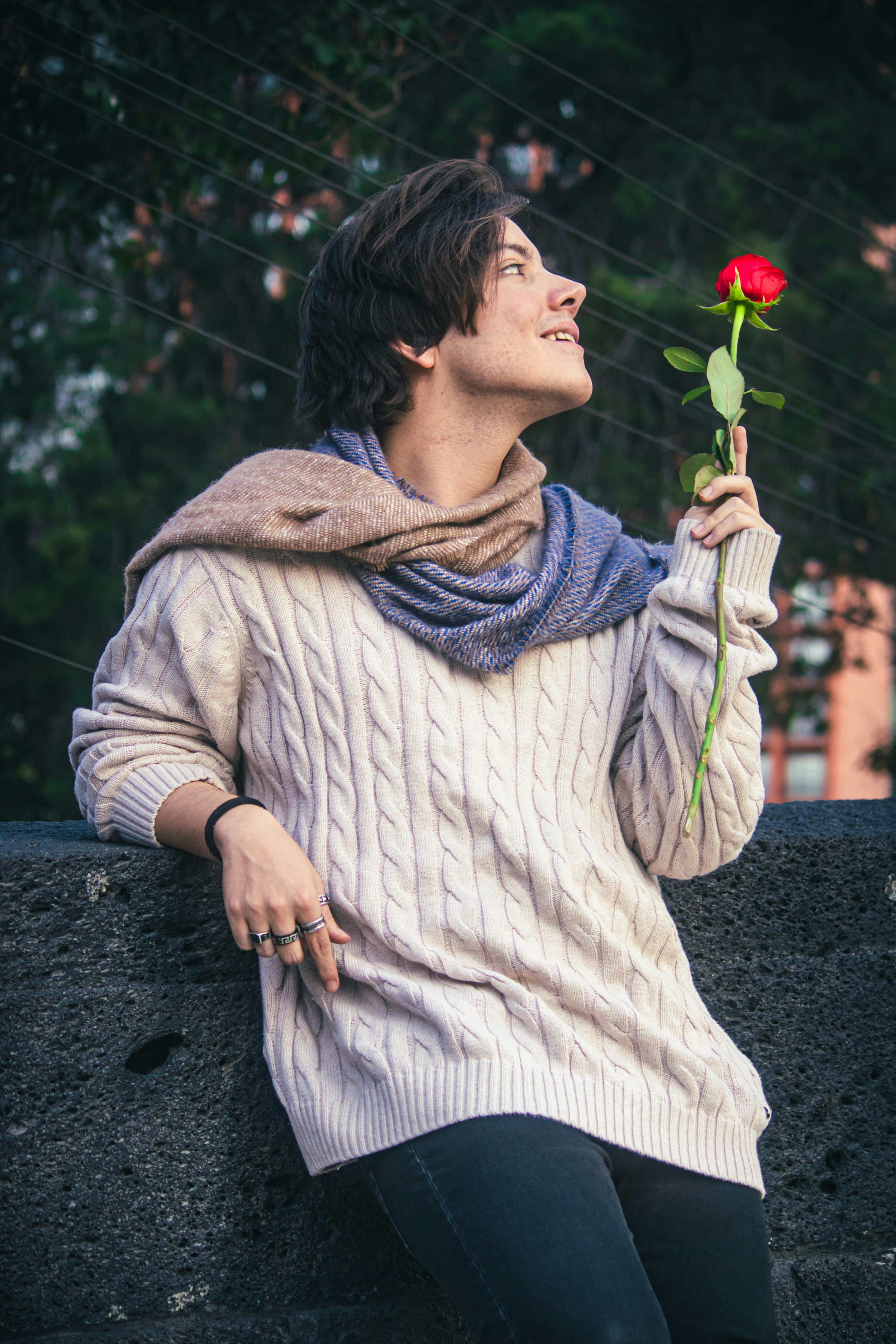 Young man in cozy sweater holding a red rose, creating a romantic vibe in an outdoor park setting.