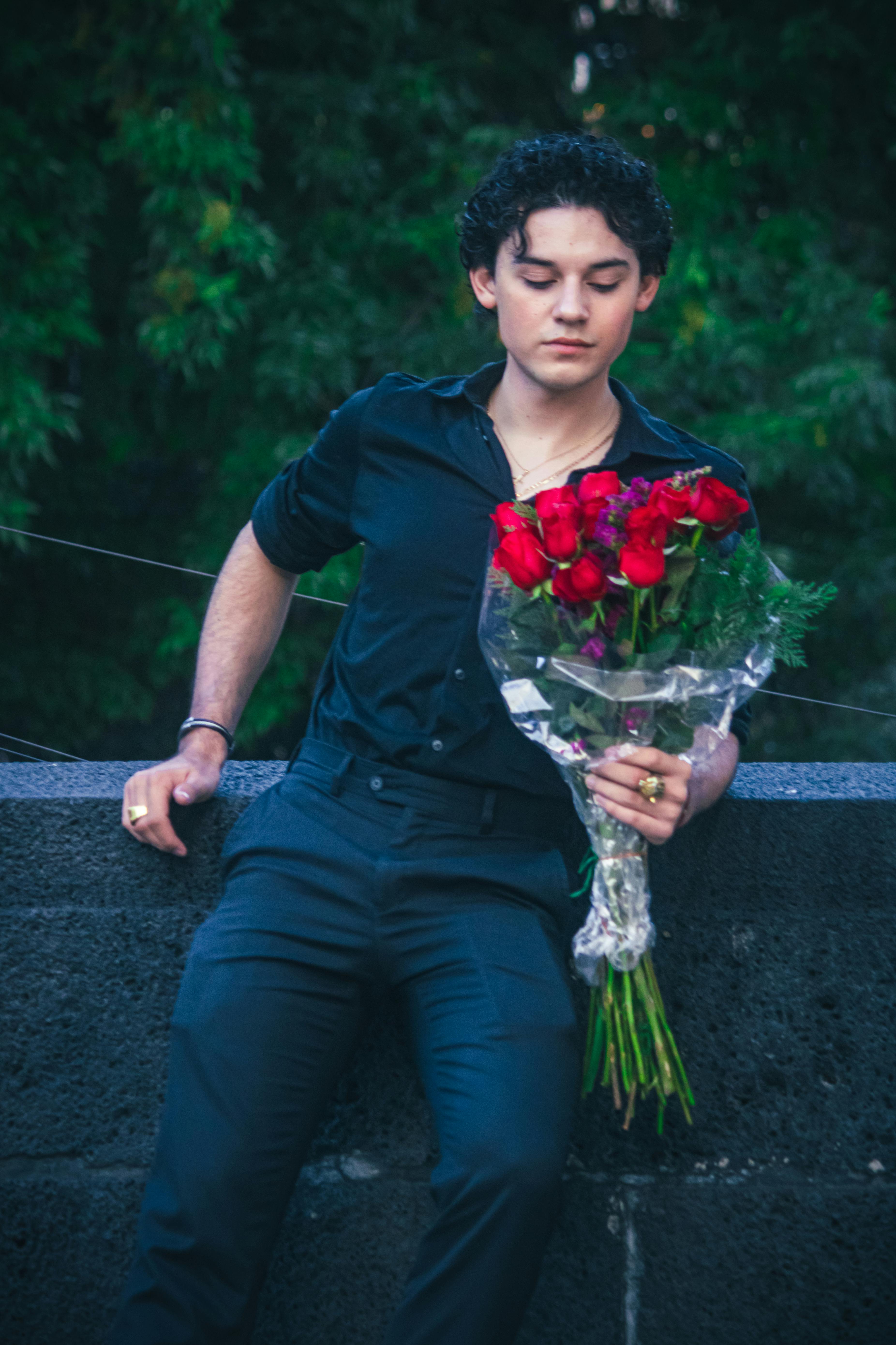 Young man in black attire holding a bouquet of red roses against a lush green backdrop, evoking romantic vibes.