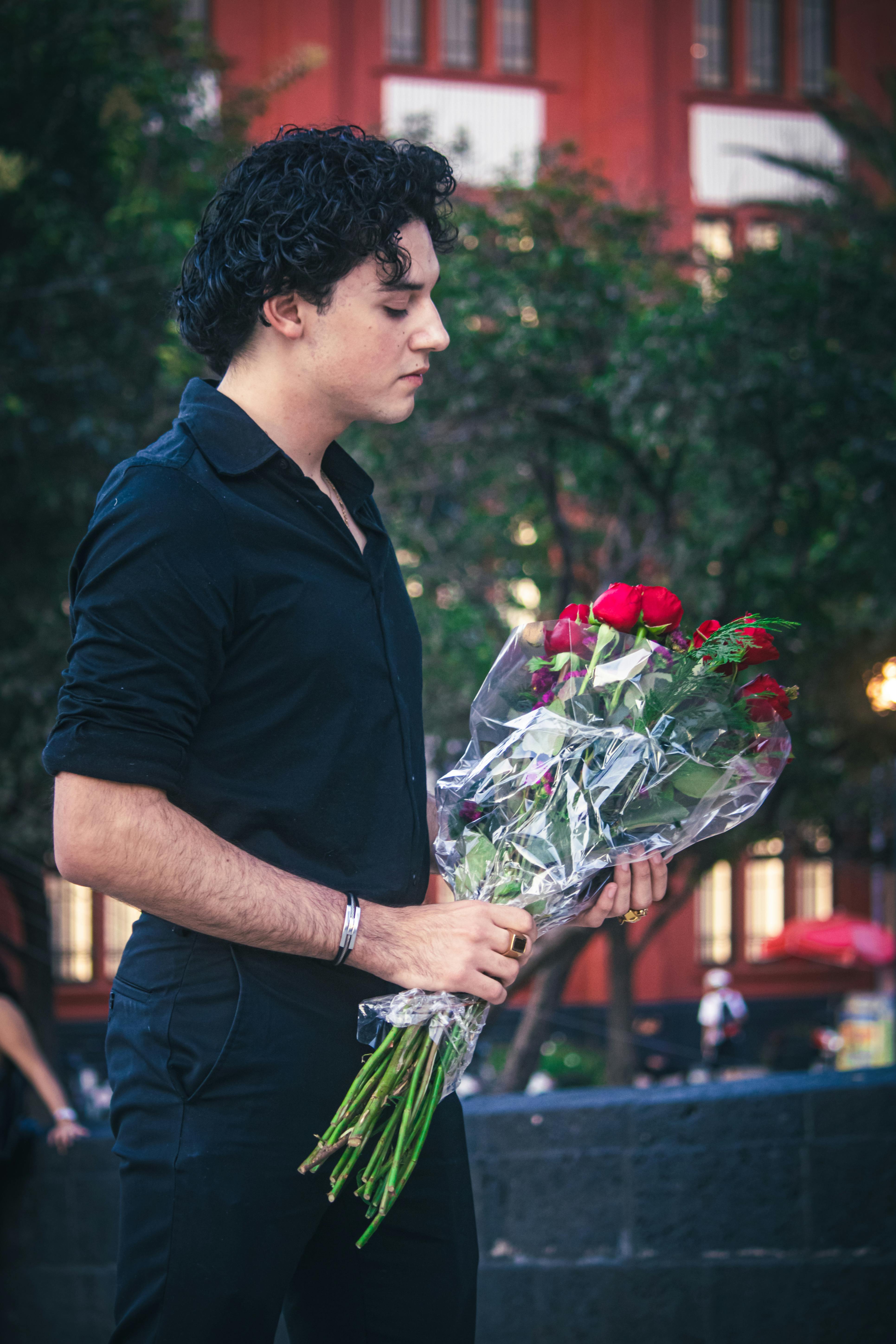 A man in black clothing holds a bouquet of red roses in a city park, conveying romance.