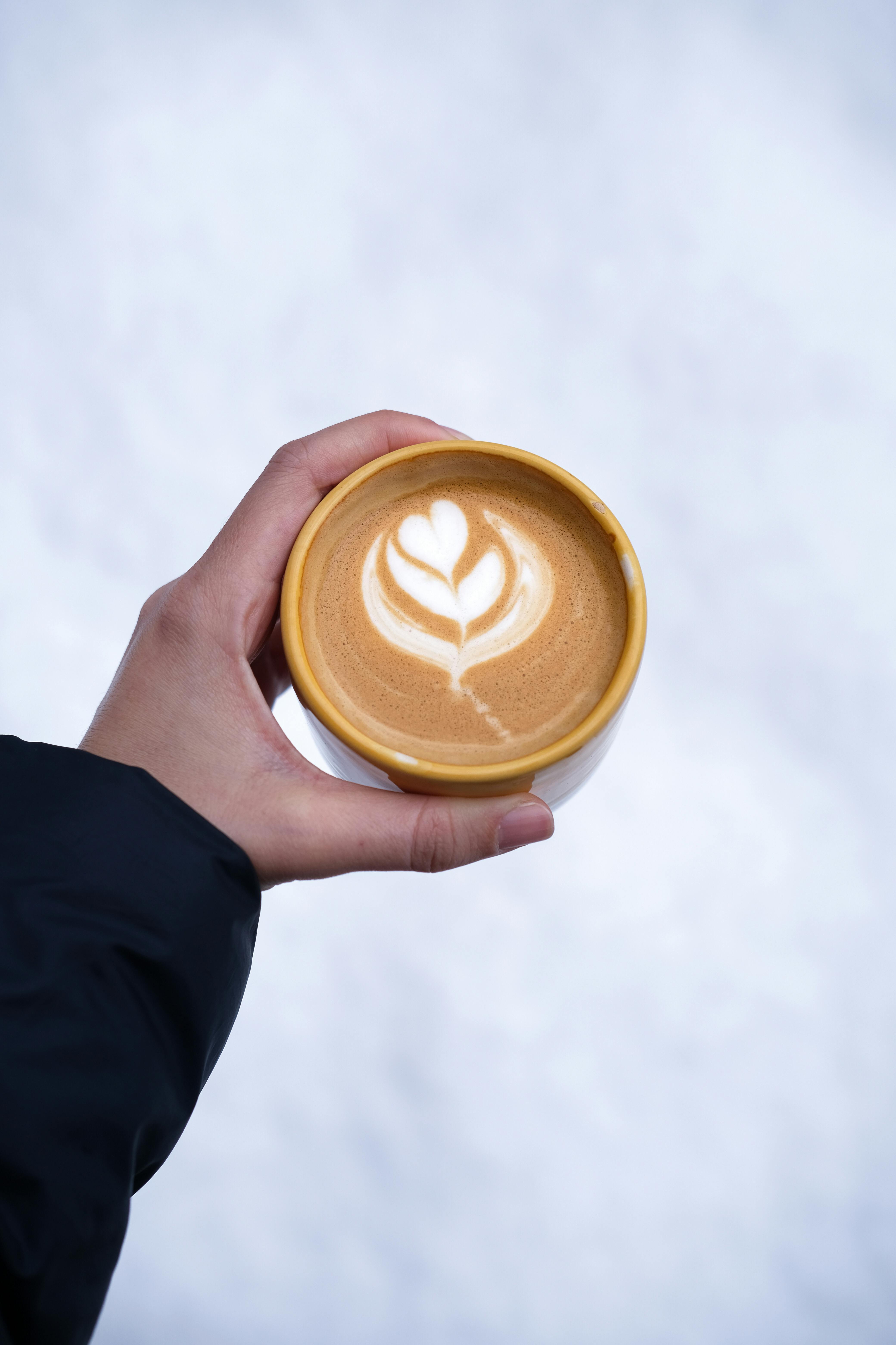 Top view of a latte with artistic foam in a yellow ceramic cup held outdoors against a snowy background.