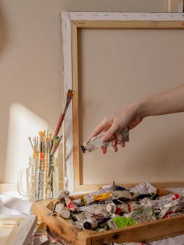 A hand reaching for paint tube in art studio with brushes and canvas frame.
