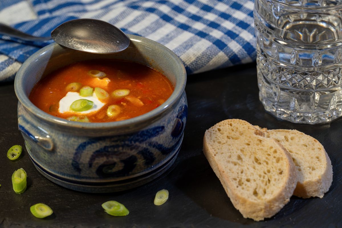 Tomato vegetable soup with sour cream and scallions in a blue stoneware crock, bread slices beside