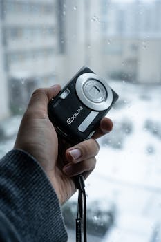 A hand holding a vintage digital camera against a snowy window background.