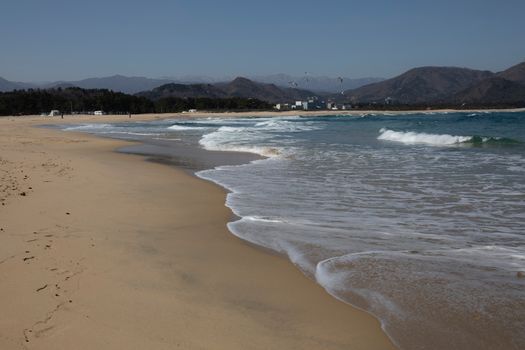 Quiet sandy beach with gentle waves and distant hills under clear blue skies, ideal for solitude seekers.