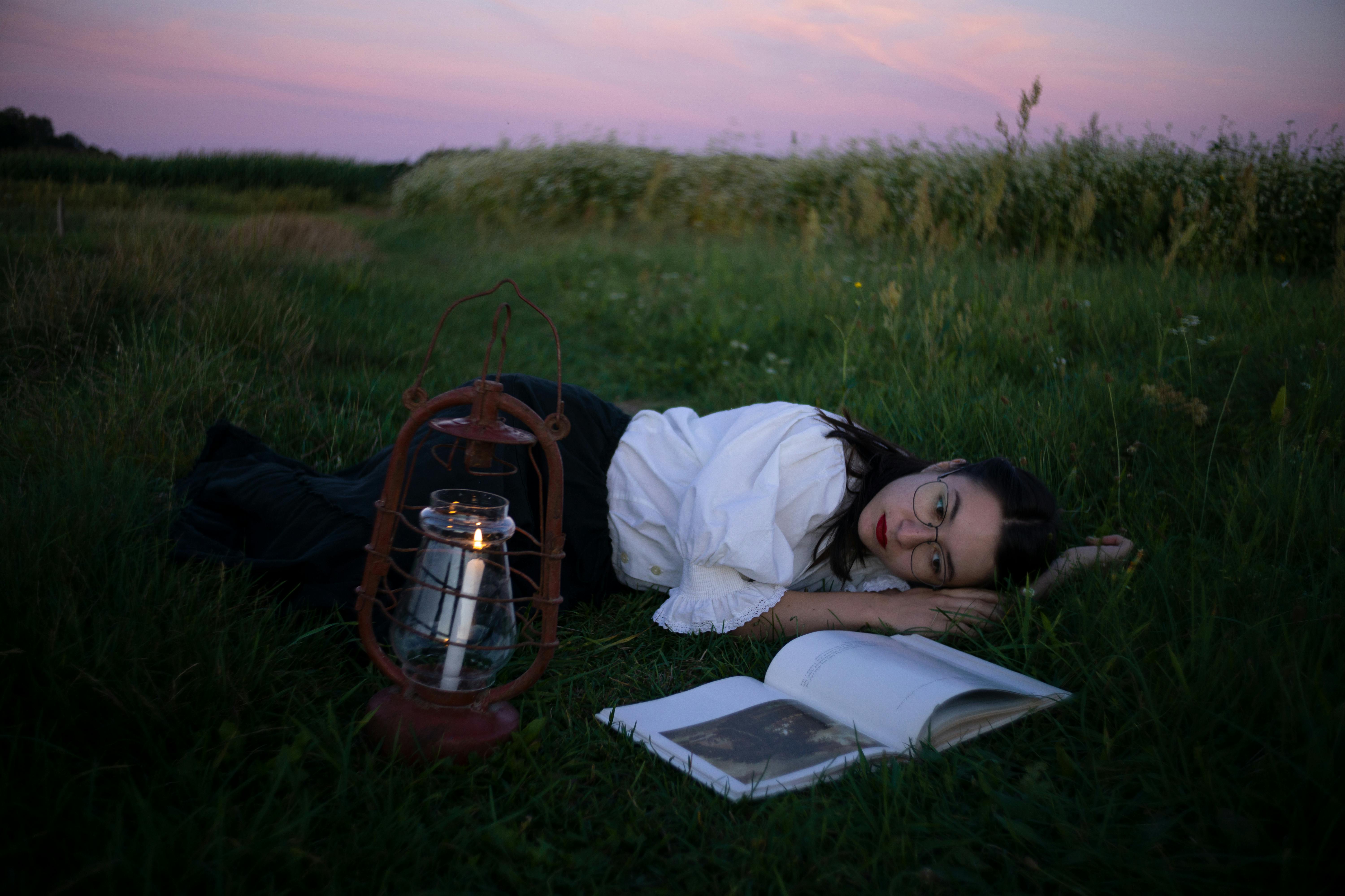 Woman relaxing with a book and lantern in a tranquil field at twilight, offering a serene escape.