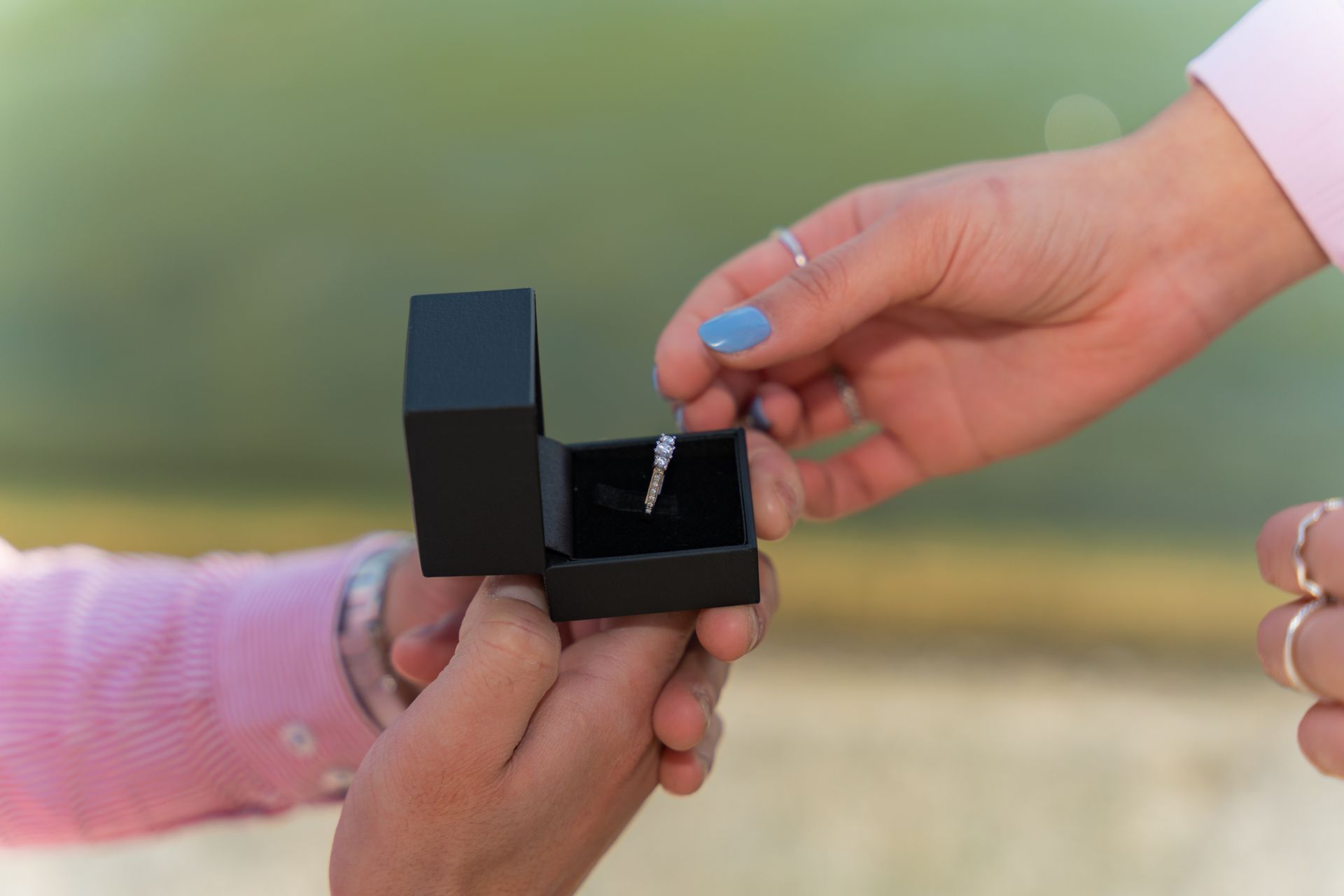 Close-up of hands exchanging a diamond ring in a black box during a proposal outdoors.