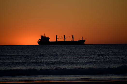 A silhouette of a large cargo ship sails against a vibrant sunset over the ocean.