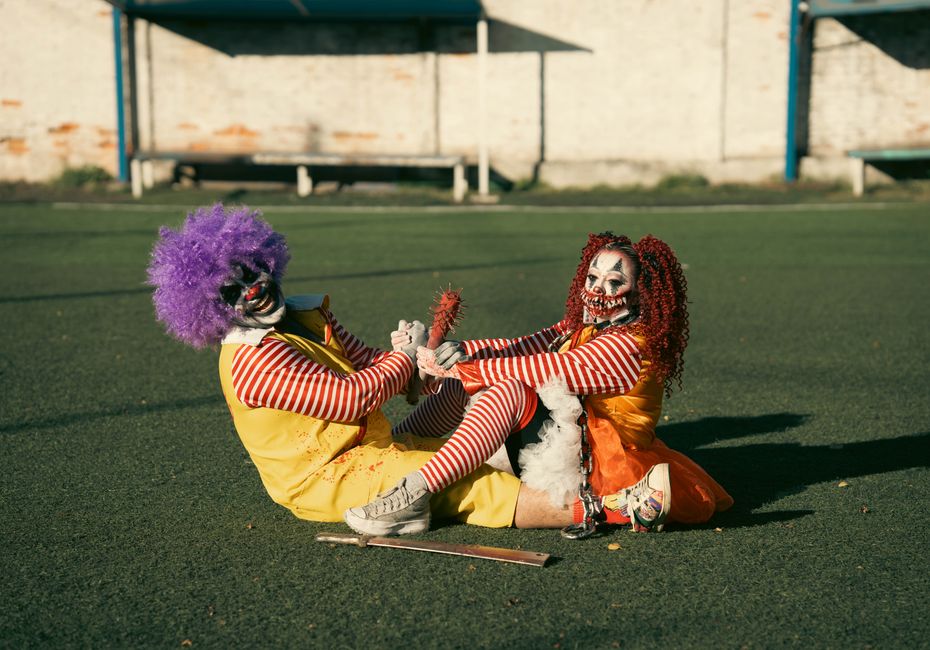Two clowns in vibrant costumes and makeup playing on a soccer field in daylight.