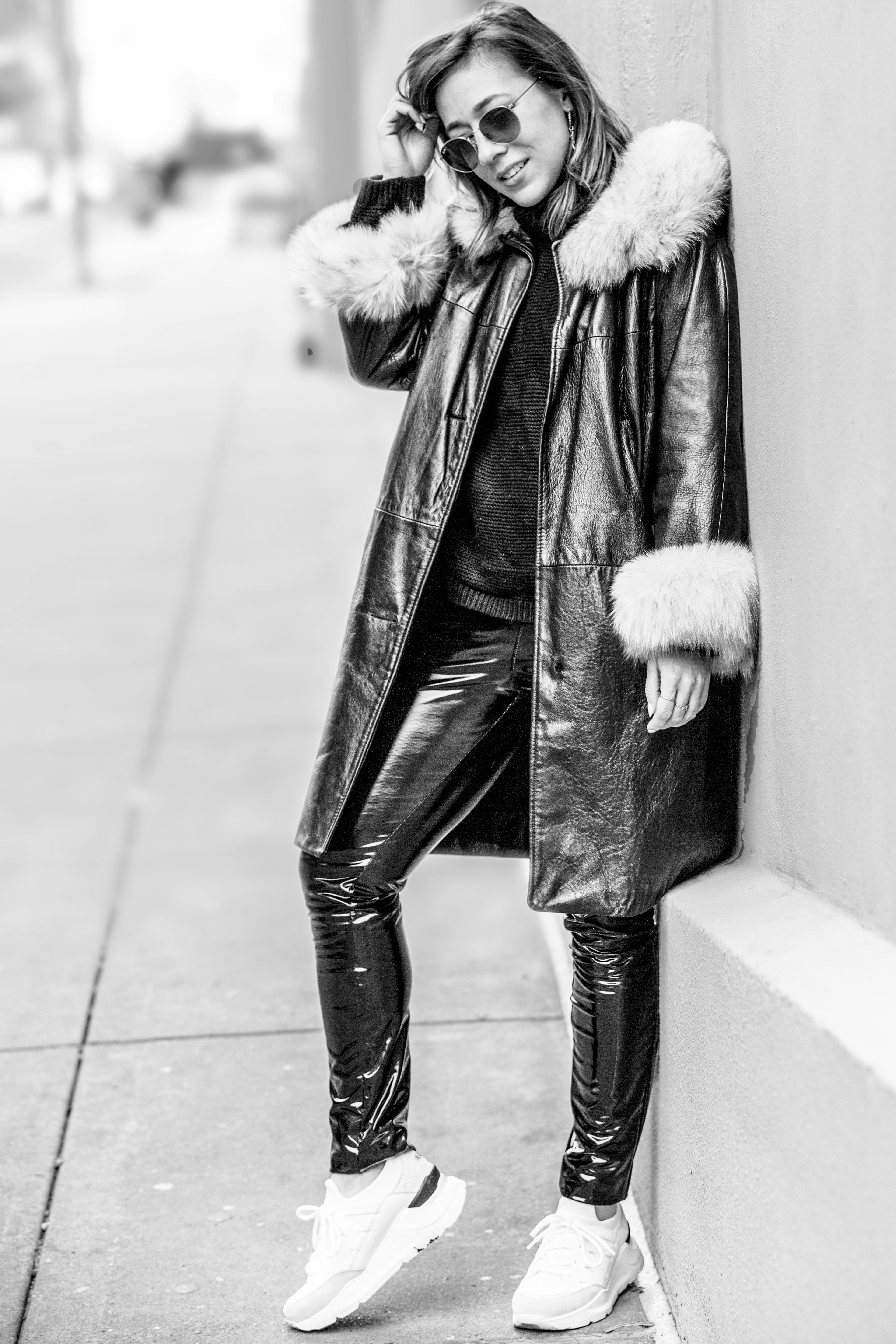 一张黑白图像中，一位时尚女性身着毛皮衬里外套和皮裤，站在户外