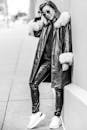一张黑白图像中，一位时尚女性身着毛皮衬里外套和皮裤，站在户外