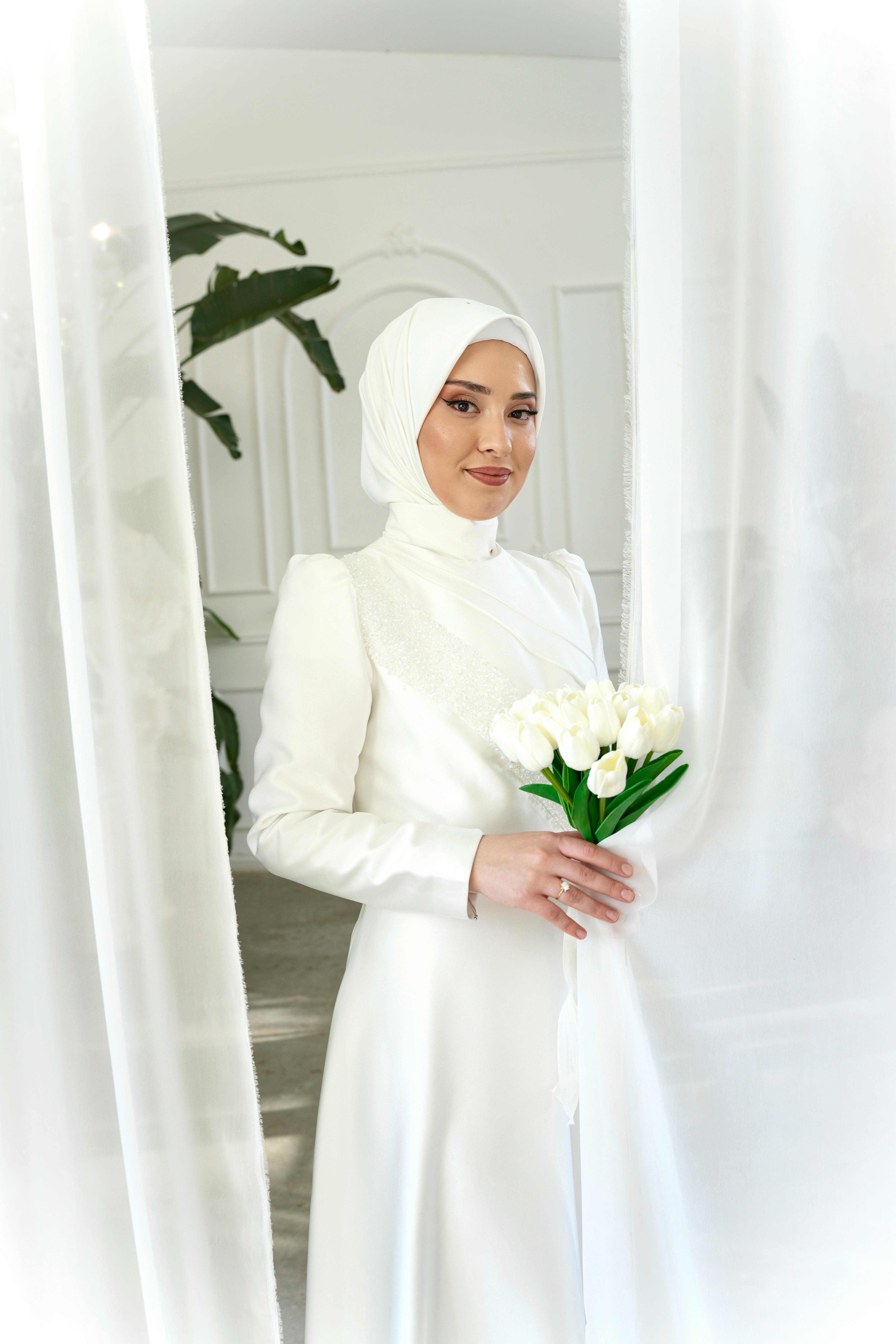 A beautiful bride in a white hijab holding white tulips indoors, exuding elegance.