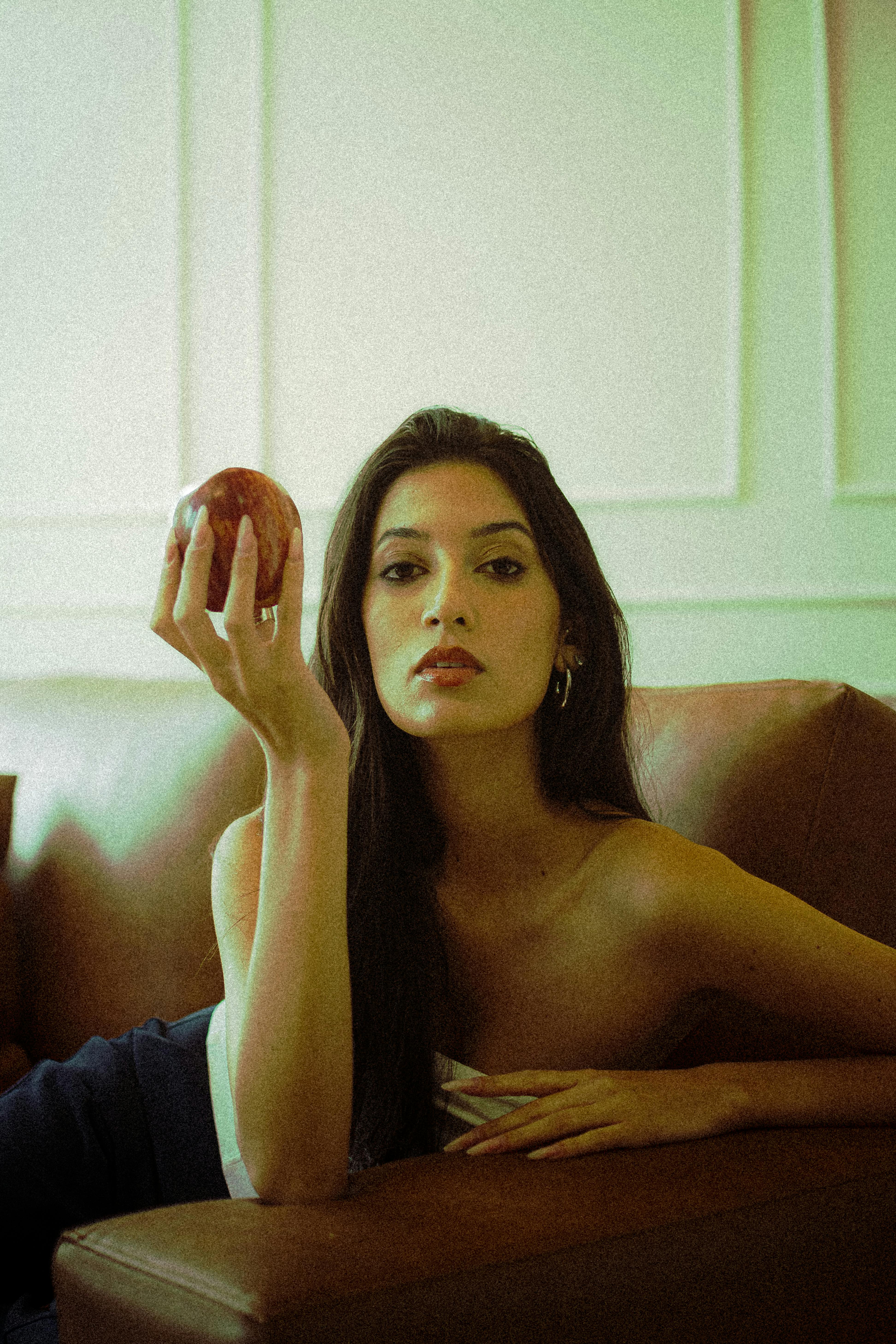 Elegant woman posing with apple on leather sofa, exuding classic allure and timeless style.