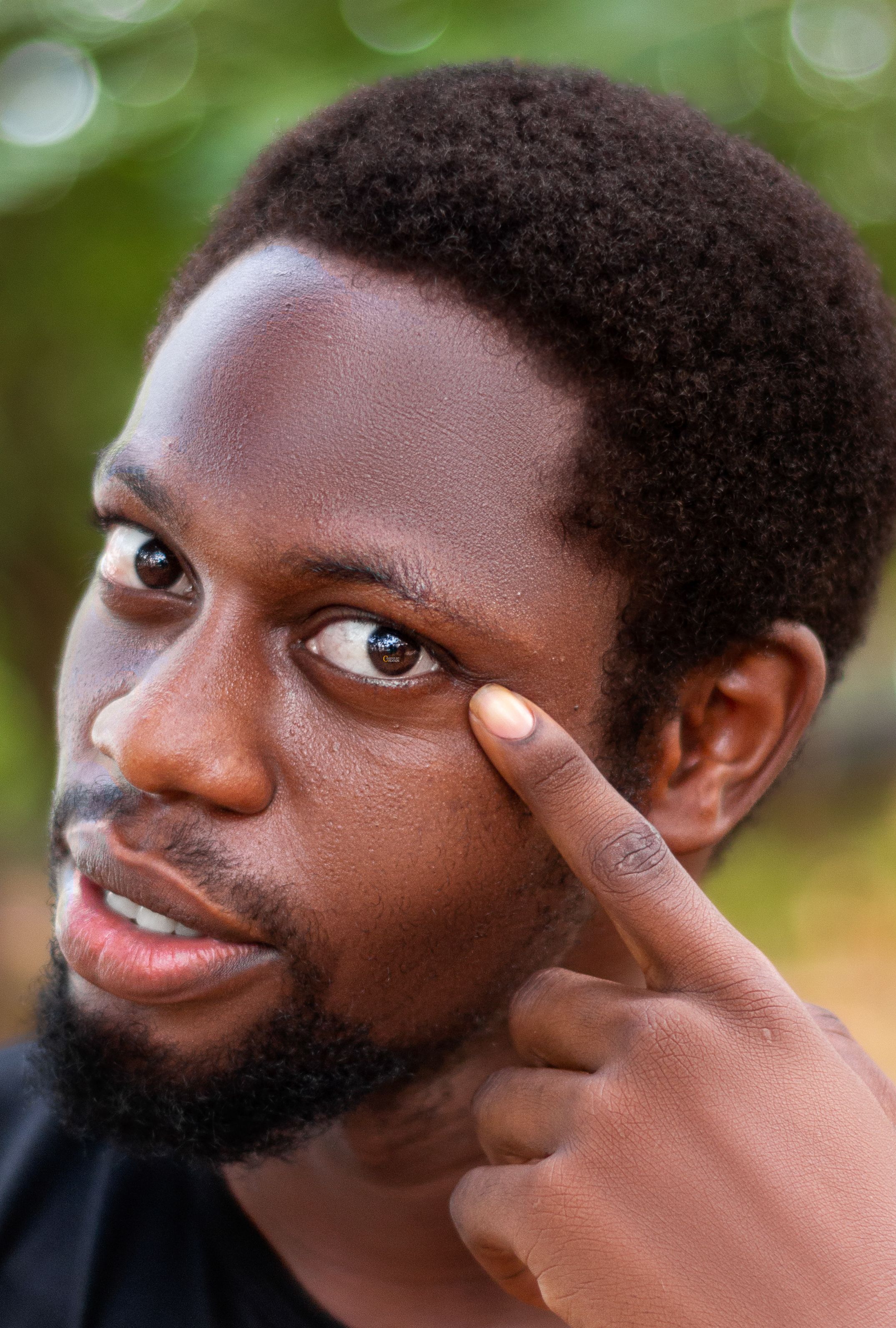 A detailed portrait showing a man pointing at his eye against a blurred outdoor background.