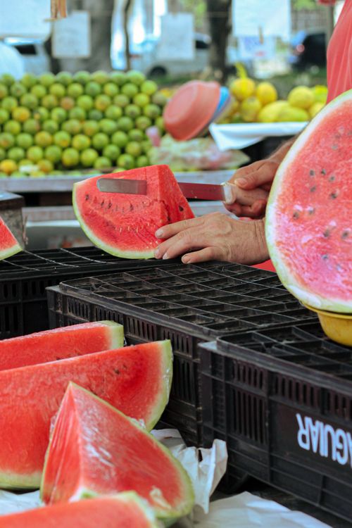 Free Fresh watermelon slices being prepared at an outdoor street market with various colorful fruits. Stock Photo