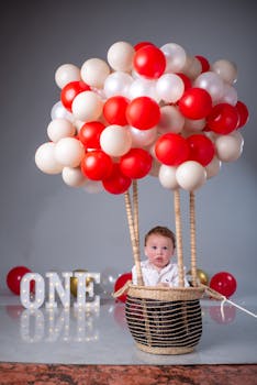Cute baby in a balloon basket celebrates first birthday with 'ONE' sign. Perfect for children's milestone moments.