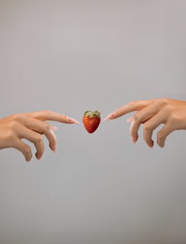 Two hands reaching towards a floating strawberry against a neutral background.