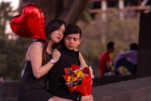 Free A couple sits close with a red heart-shaped balloon and a bouquet, capturing romance. Stock Photo
