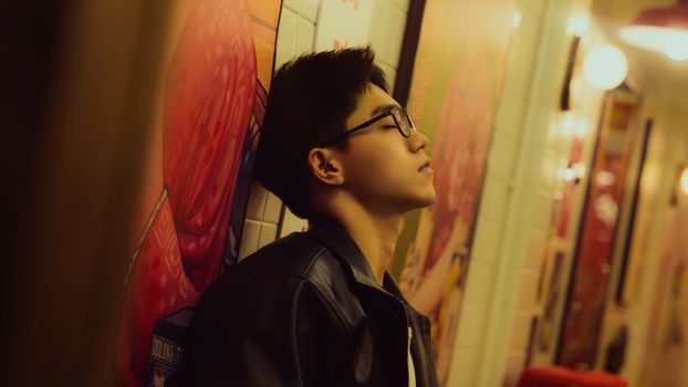 A man in a leather jacket leans against a colorful wall, lost in thought.