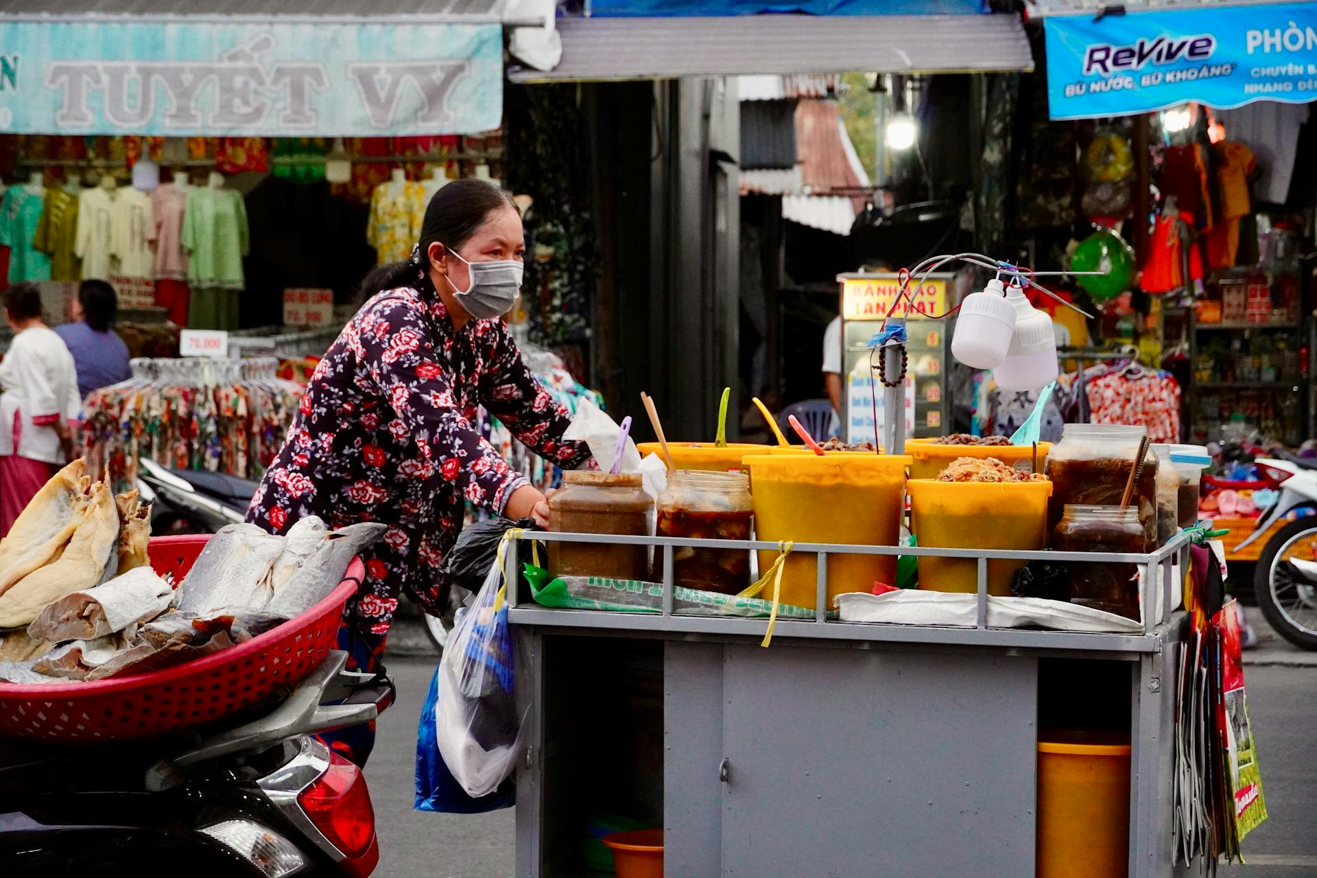 A masked female vendor pushes a colorful food cart in a bustling street market setting.